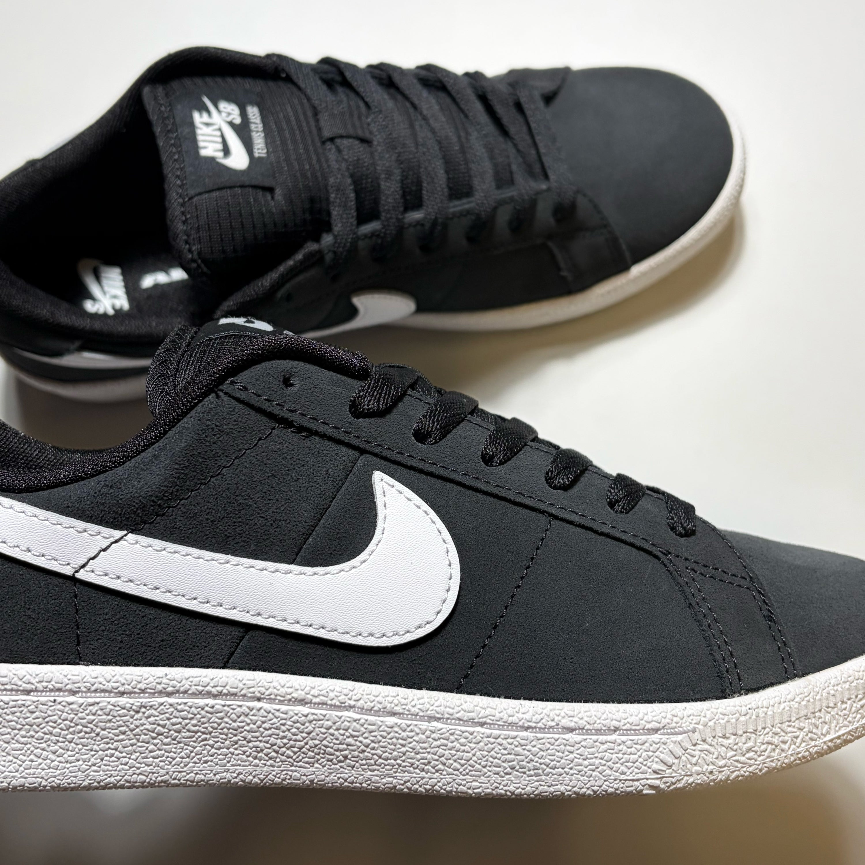 A pair of Nike SB Tennis Classic sneakers in black and white, featuring a black suede upper, black laces, and a prominent white Swoosh on the side, set against a plain light background.