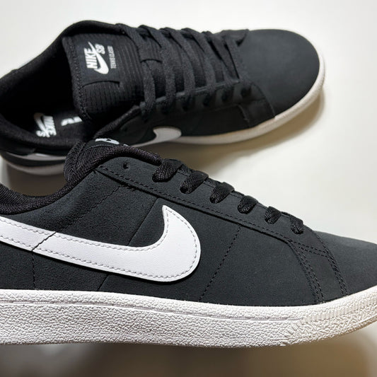 A pair of Nike SB Tennis Classic sneakers in black and white, featuring a black suede upper, black laces, and a prominent white Swoosh on the side, set against a plain light background.