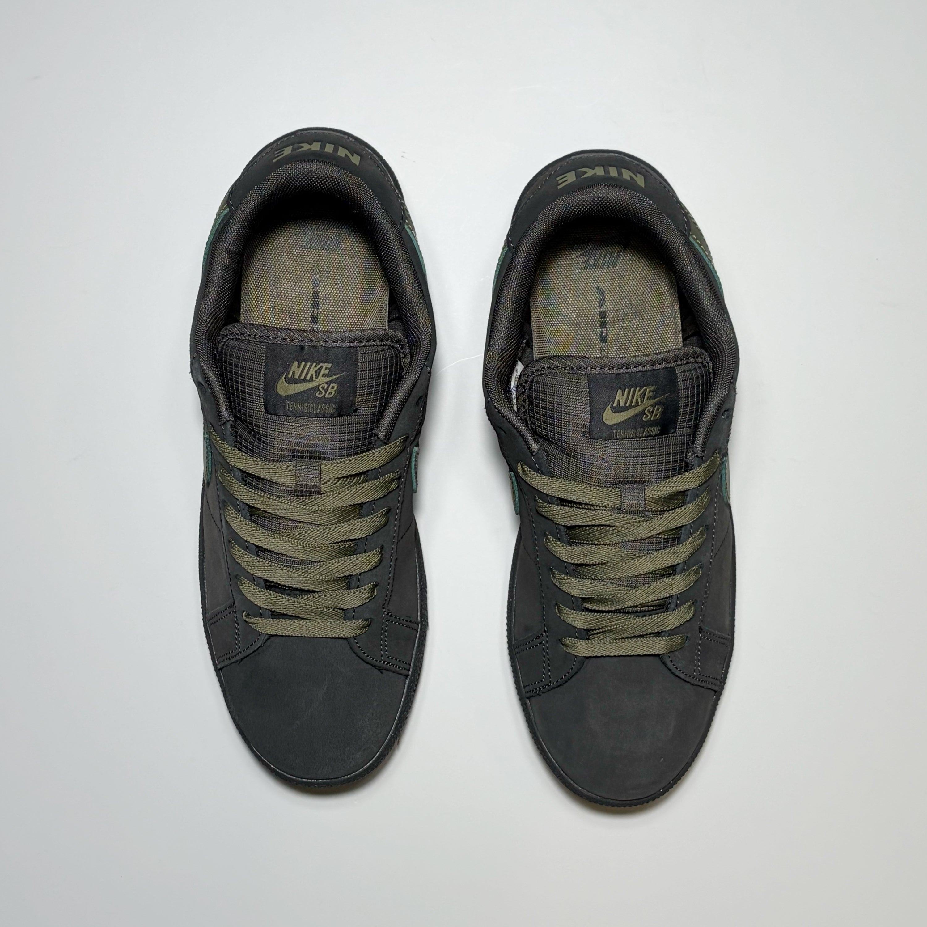 A pair of Nike SB sneakers in dark gray suede and mesh with olive green laces, shown from a top-down view on a light gray background.