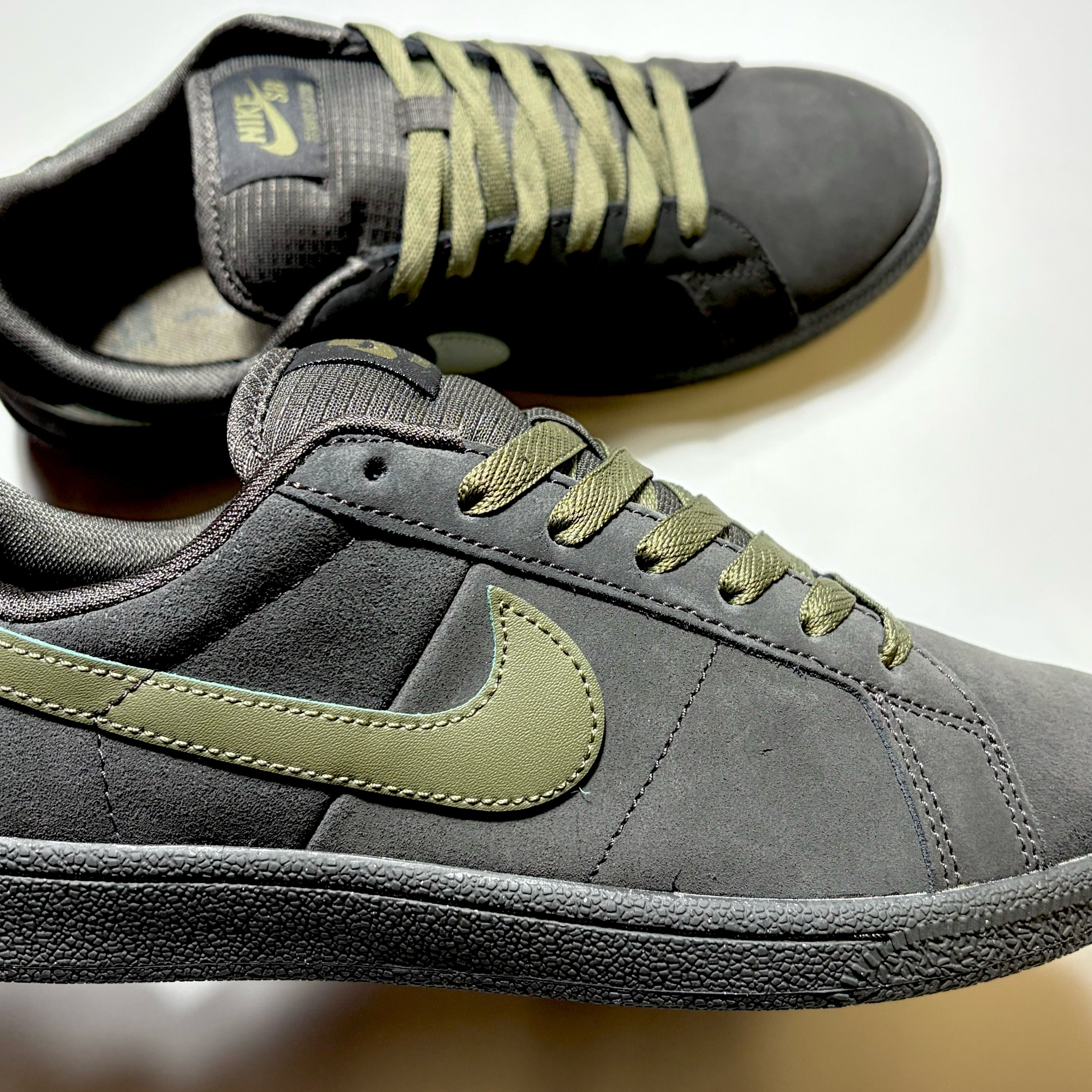 A pair of Nike SB low-top sneakers in dark gray suede with olive green laces and a matching olive green Swoosh logo, shown on a plain light background.