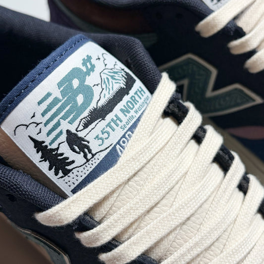 Close-up of a New Balance sneaker showing the tongue label with '35th North' graphics, cream laces, and a dark navy suede upper.