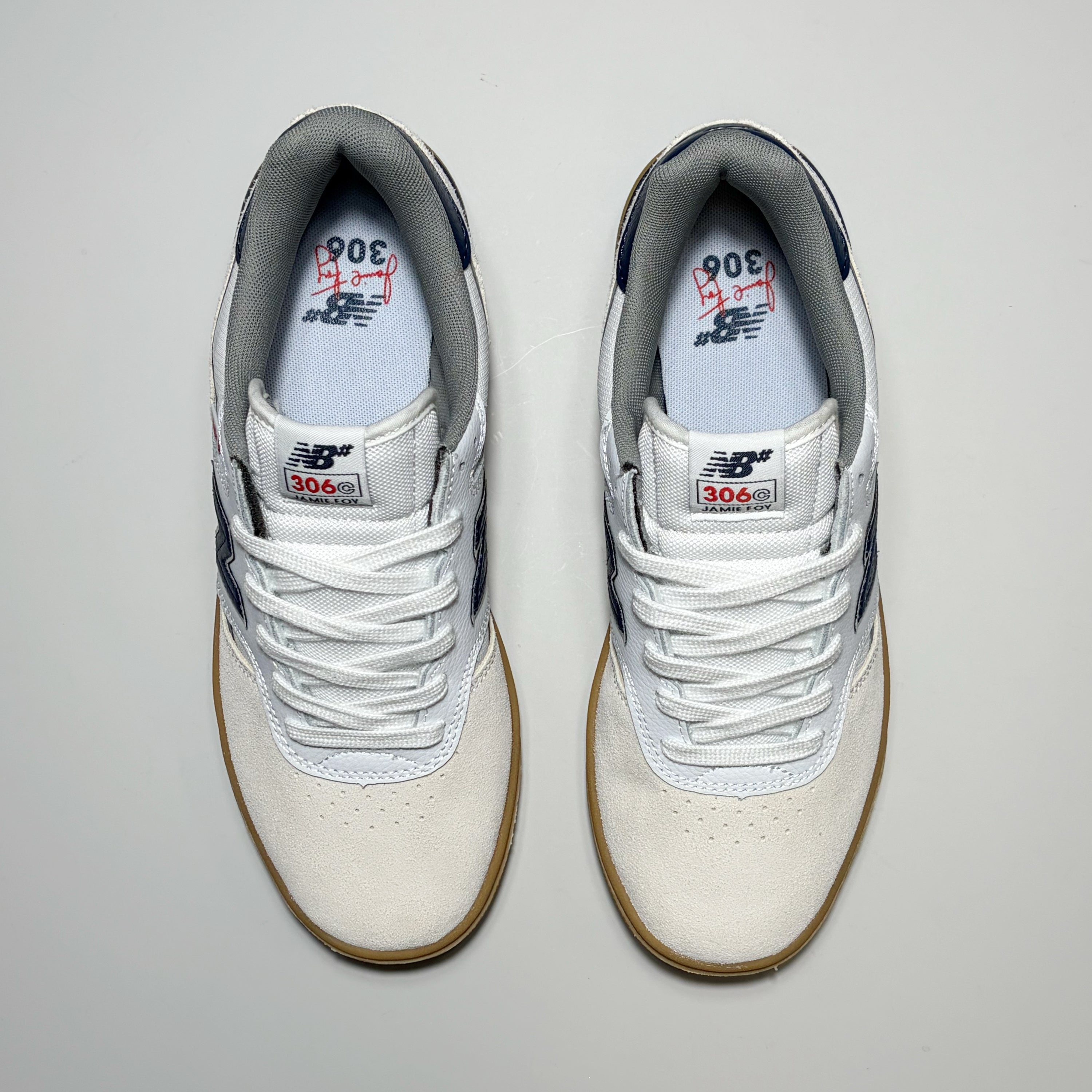 A pair of New Balance Numeric NM306C sneakers shown from a top-down view on a light gray background. The shoes feature an off-white suede and mesh upper, white laces, navy blue accents on the heel and tongue, and a brown gum sole.