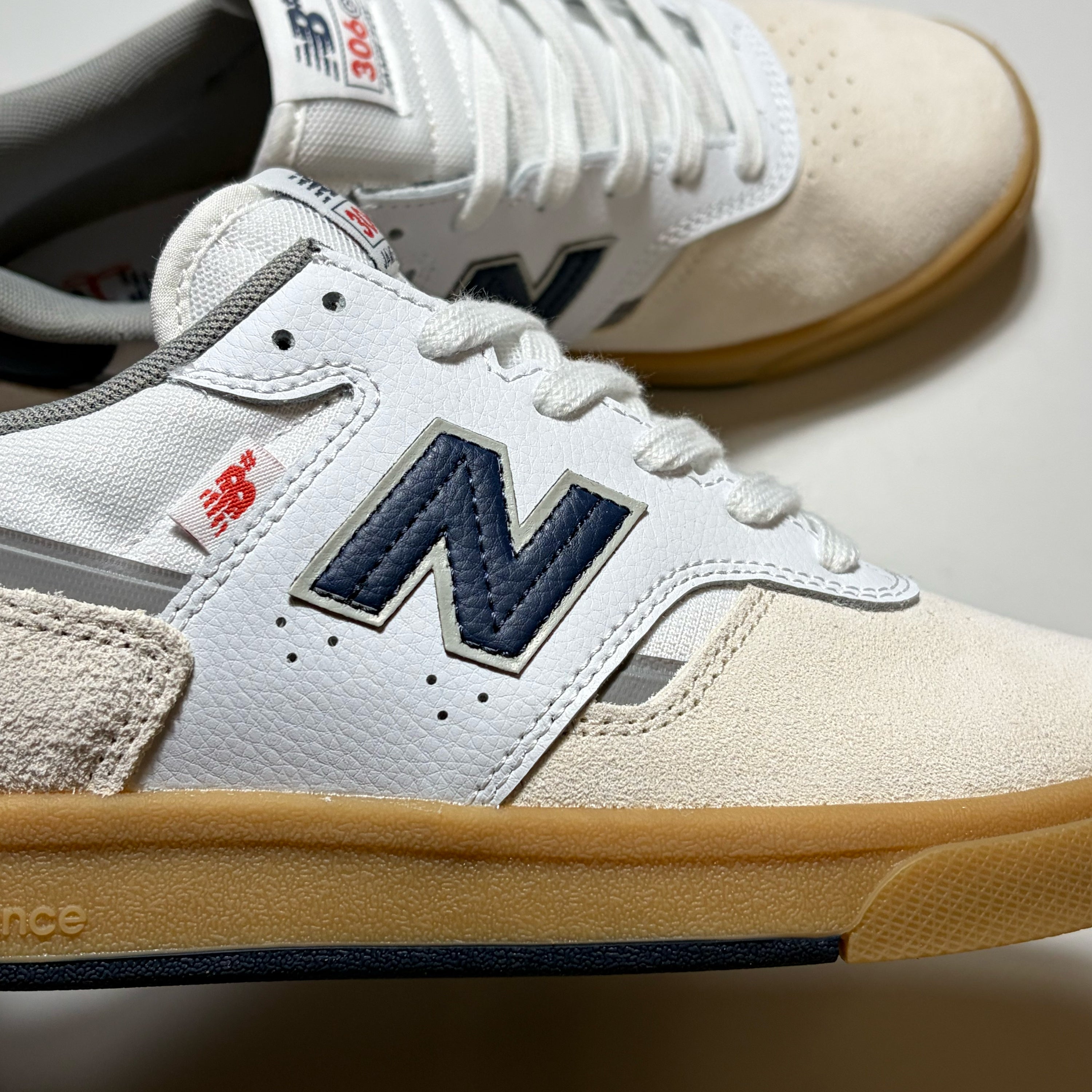 A close-up of a New Balance sneaker in off-white suede and leather with a navy blue 'N' logo, white laces, a red brand tag, and a tan gum sole, with another shoe blurred in the background.