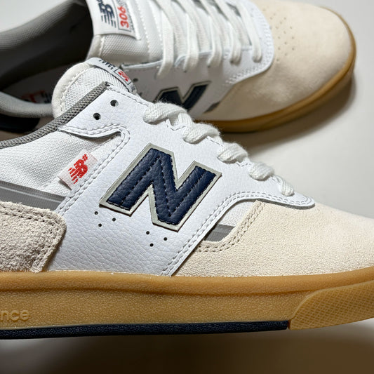 A close-up of a New Balance sneaker in off-white suede and leather with a navy blue 'N' logo, white laces, a red brand tag, and a tan gum sole, with another shoe blurred in the background.