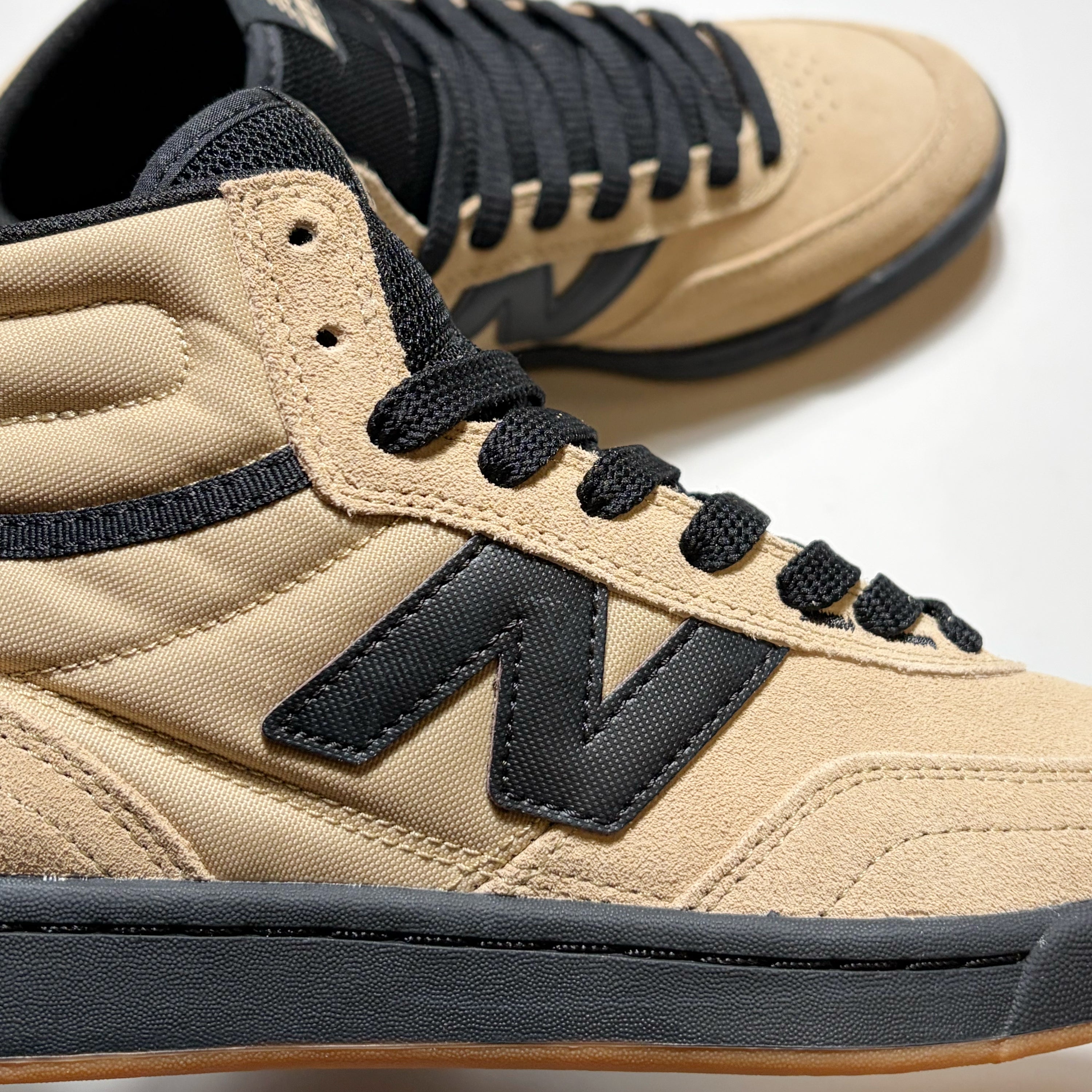 A close-up of a beige canvas high-top sneaker with black laces, a black 'N' logo, and a dark gray midsole, with a second shoe blurred in the background.