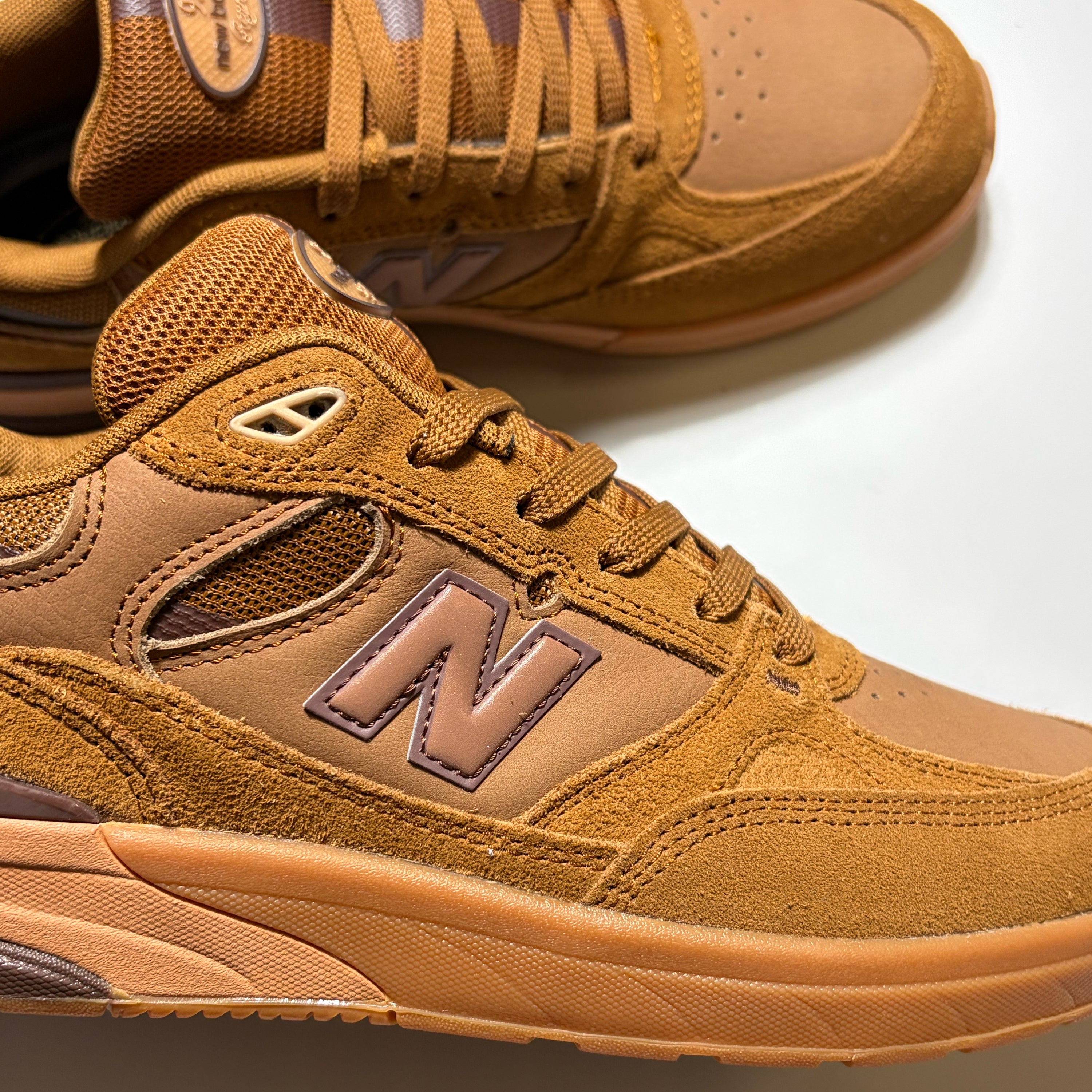 A close-up of a New Balance Numeric 933 sneaker in a warm tan and brown suede and mesh combination, with the signature 'N' logo visible on the side. Another shoe is partially visible in the background on a clean white surface.