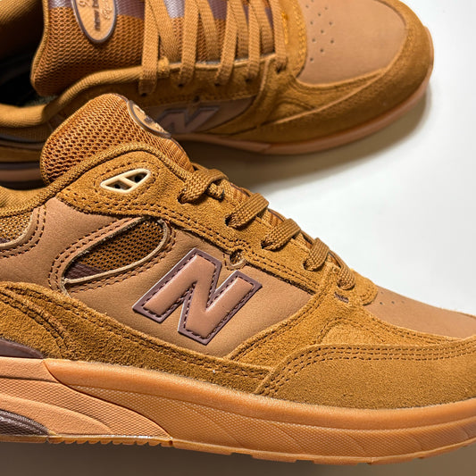 A close-up of a New Balance Numeric 933 sneaker in a warm tan and brown suede and mesh combination, with the signature 'N' logo visible on the side. Another shoe is partially visible in the background on a clean white surface.