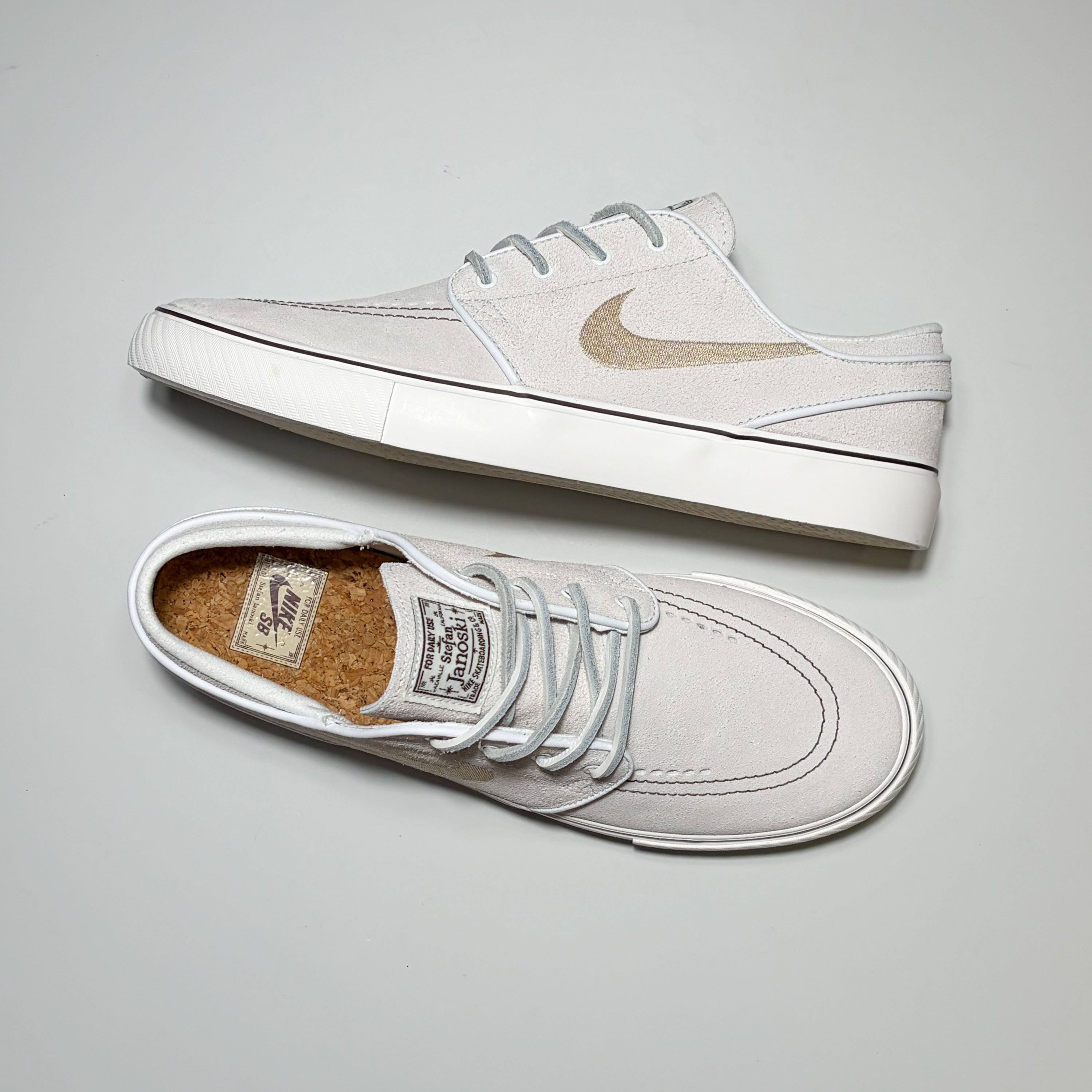 A pair of Nike SB Janoski sneakers in off-white suede with light gray laces and a metallic gold Swoosh, displayed on a neutral light gray background.