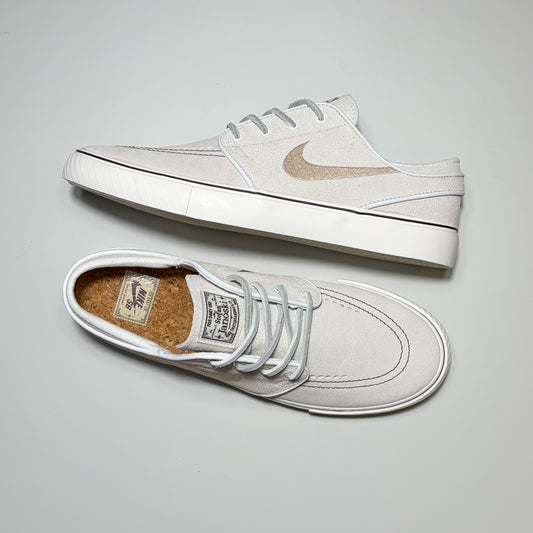 A pair of Nike SB Janoski sneakers in off-white suede with light gray laces and a metallic gold Swoosh, displayed on a neutral light gray background.