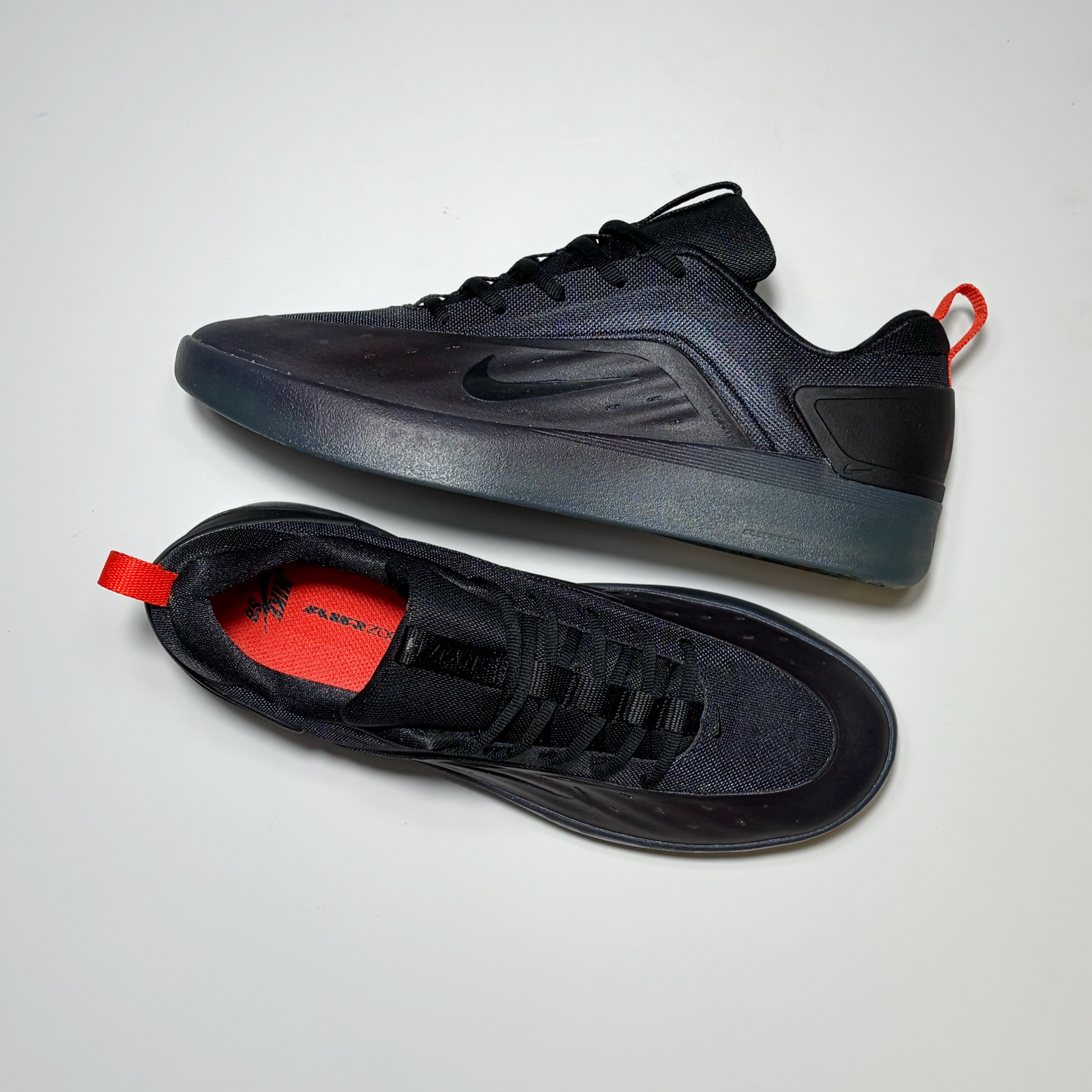 A pair of Nike SB Nyjah 4 sneakers in black, shown from both the side and top-down views on a light gray background. The shoes feature a black mesh and synthetic upper, a translucent dark gray midsole, and a bright red pull tab and interior lining.