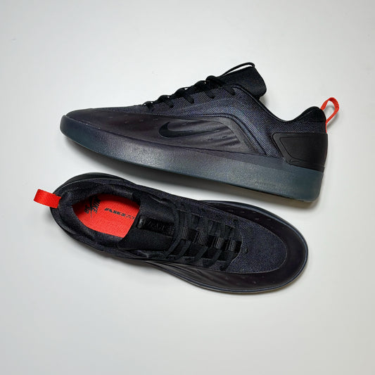 A pair of Nike SB Nyjah 4 sneakers in black, shown from both the side and top-down views on a light gray background. The shoes feature a black mesh and synthetic upper, a translucent dark gray midsole, and a bright red pull tab and interior lining.