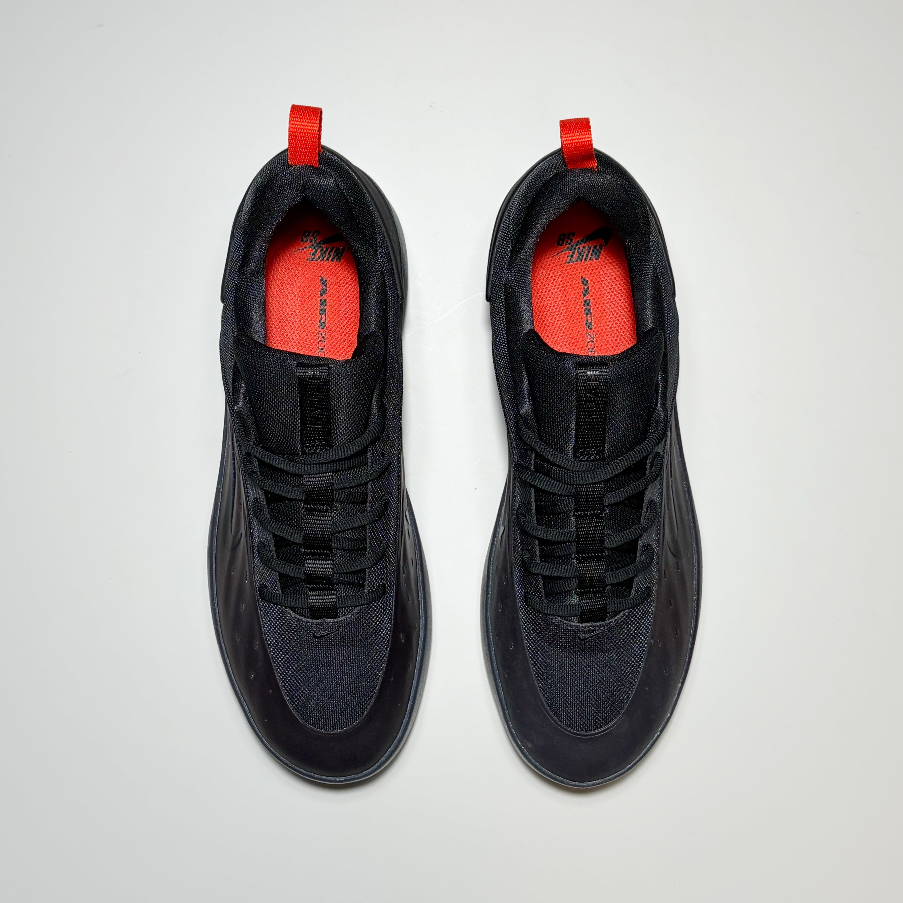 A pair of Nike SB Nyjah 4 shoes shown from a top-down view. The shoes are primarily black with a textured mesh upper, black laces, and bright red pull tabs and lining.