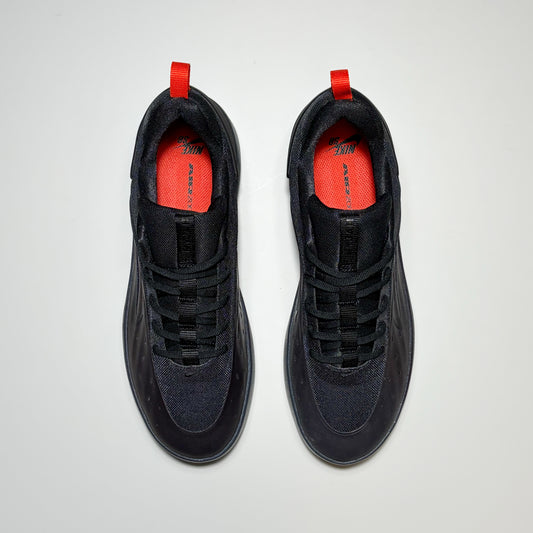 A pair of Nike SB Nyjah 4 shoes shown from a top-down view. The shoes are primarily black with a textured mesh upper, black laces, and bright red pull tabs and lining.
