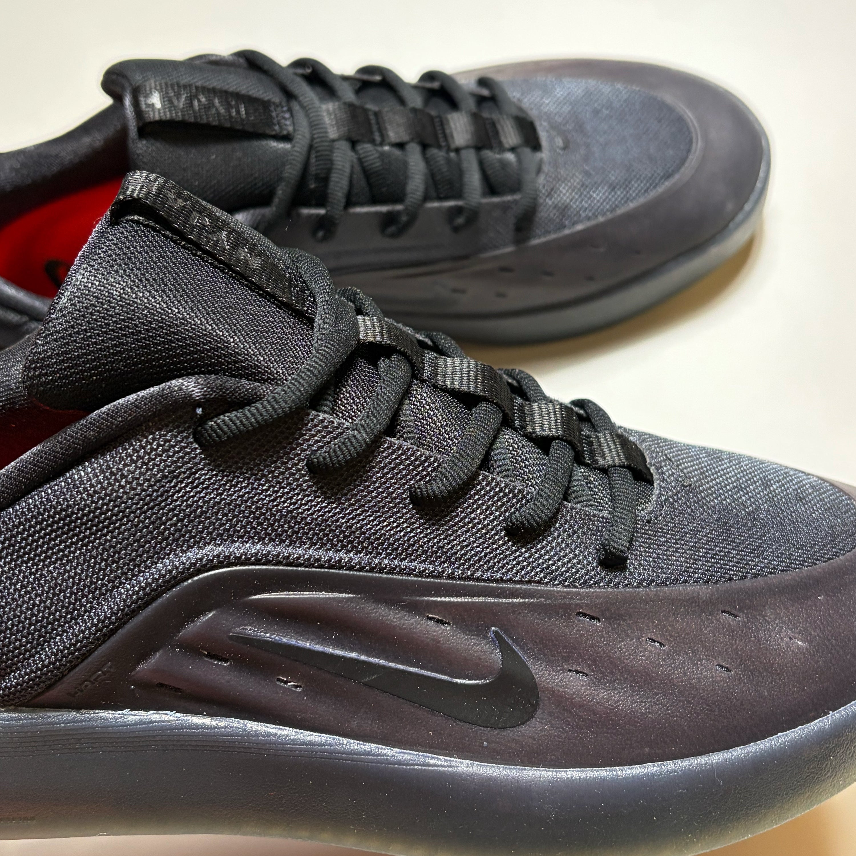 A close-up of a black Nike SB Nyjah  sneaker with a textured mesh upper, black laces, and a black Swoosh on the side, with a second matching shoe blurred in the background.