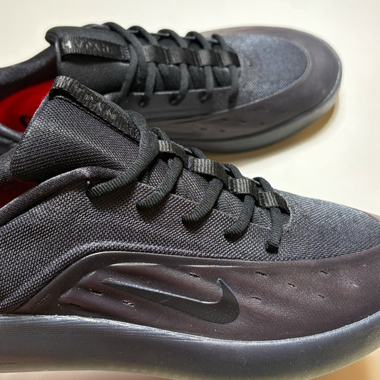 A close-up of a black Nike SB Nyjah  sneaker with a textured mesh upper, black laces, and a black Swoosh on the side, with a second matching shoe blurred in the background.