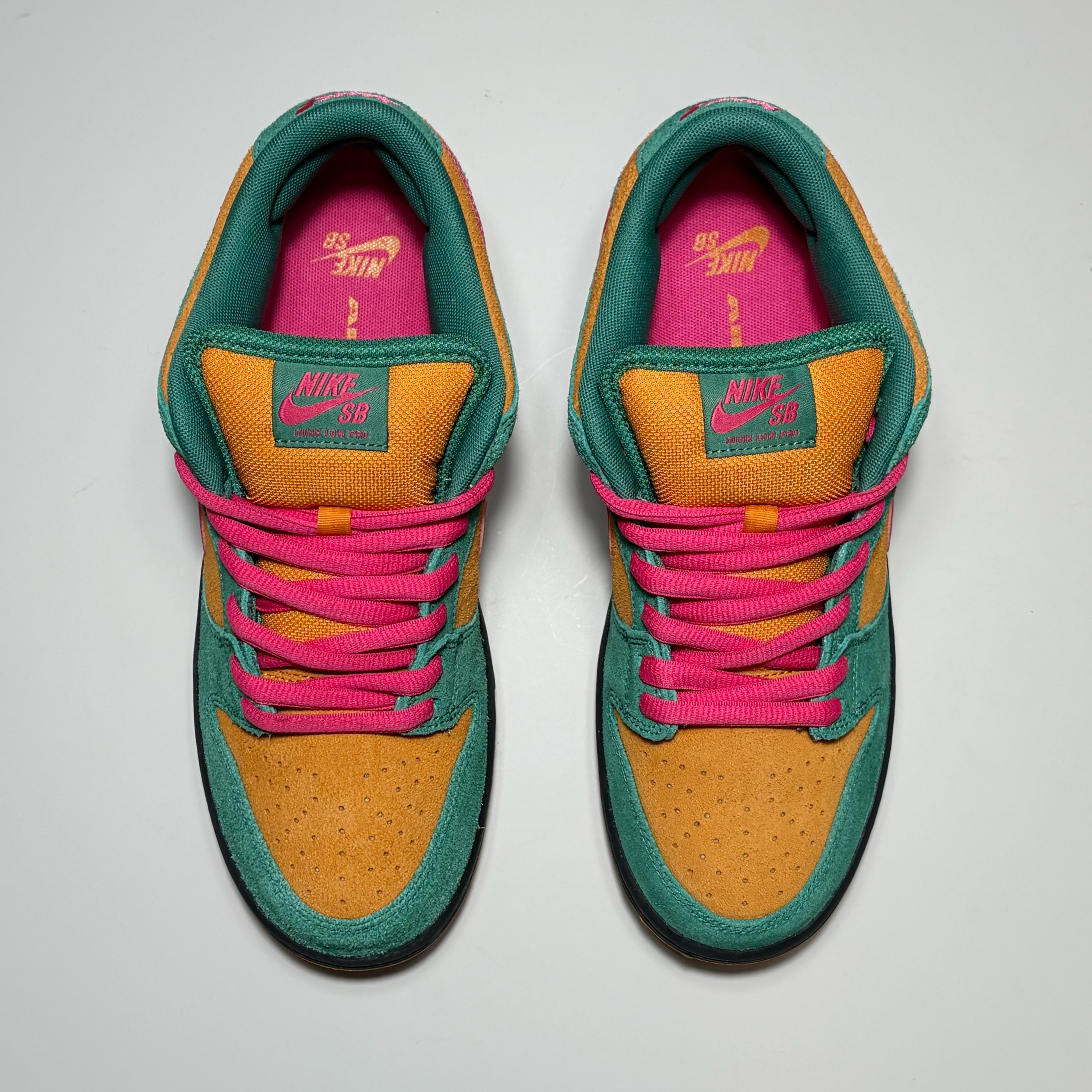 A pair of Nike SB Dunk Low sneakers shown from a top-down view on a light gray background. The shoes feature a mustard yellow suede toe box, teal green suede overlays, bright pink laces, and a vibrant pink interior lining with yellow Nike branding.