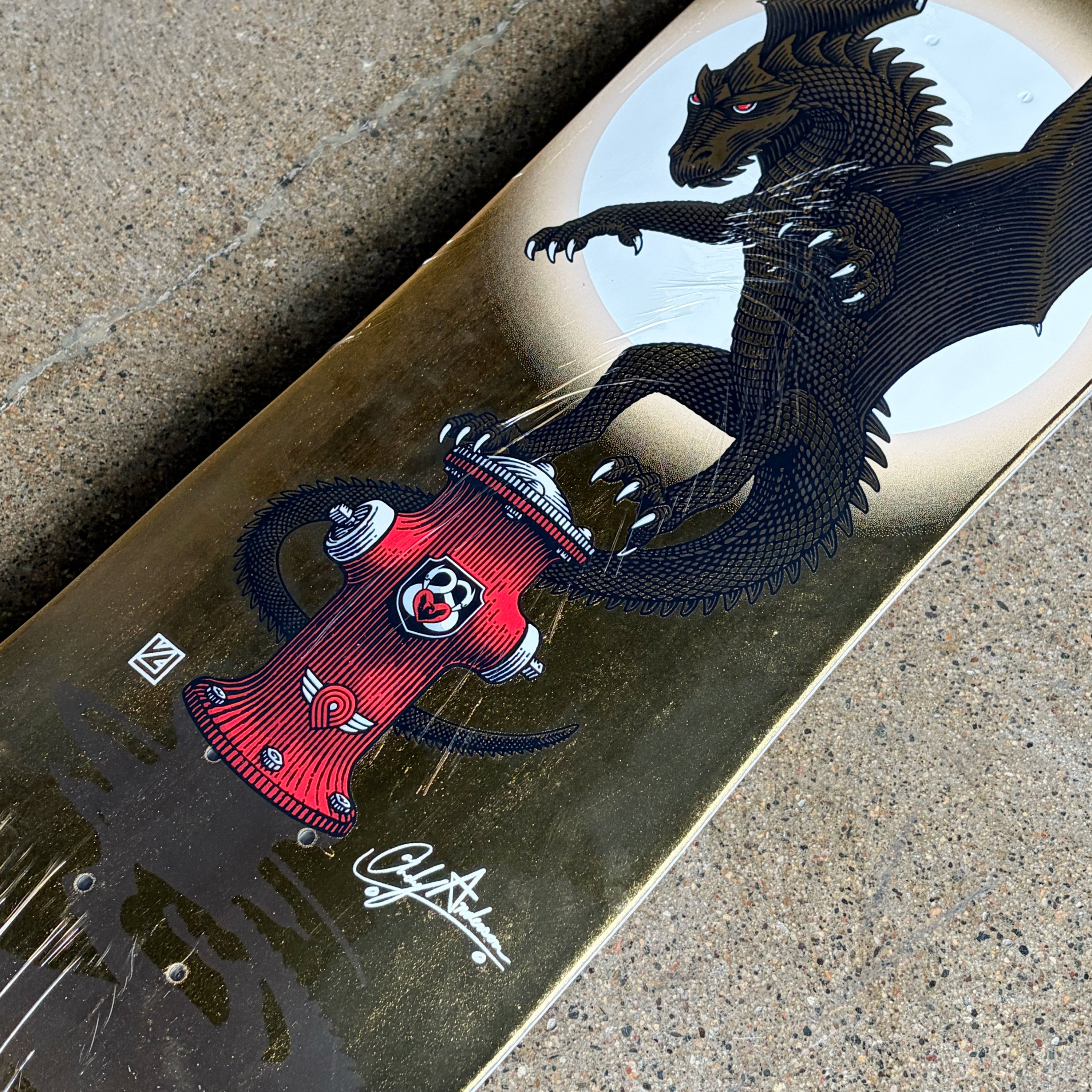 A close-up of a skateboard deck lying on concrete, showing a graphic design of a black dragon-like creature with red eyes and a red fire hydrant with a clown face, over a metallic gold background.