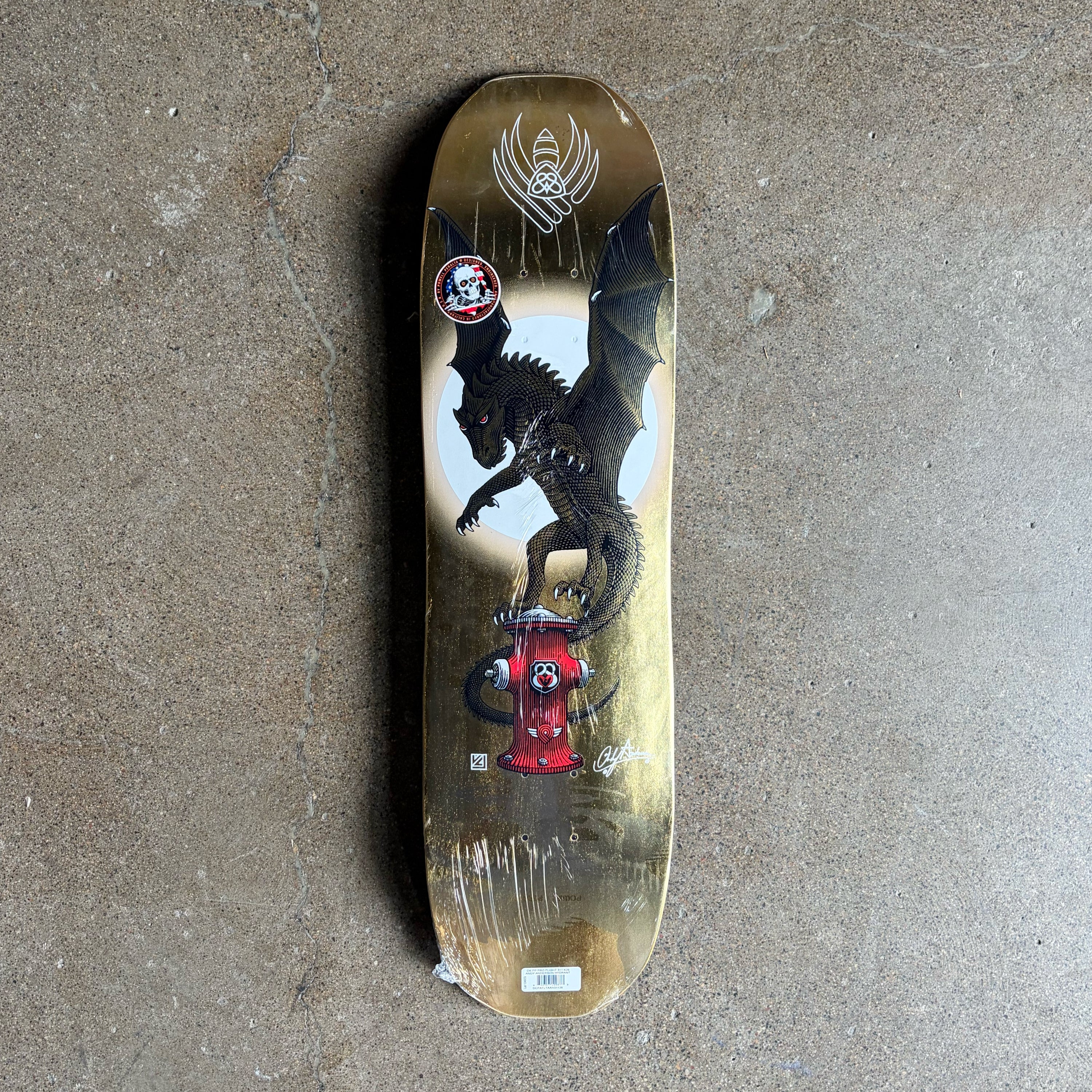 A Powell Flight Andy Anderson Hydrant deck featuring a black dragon standing on a red fire hydrant against a white circular backdrop. The deck is photographed from directly above on a textured gray concrete floor.