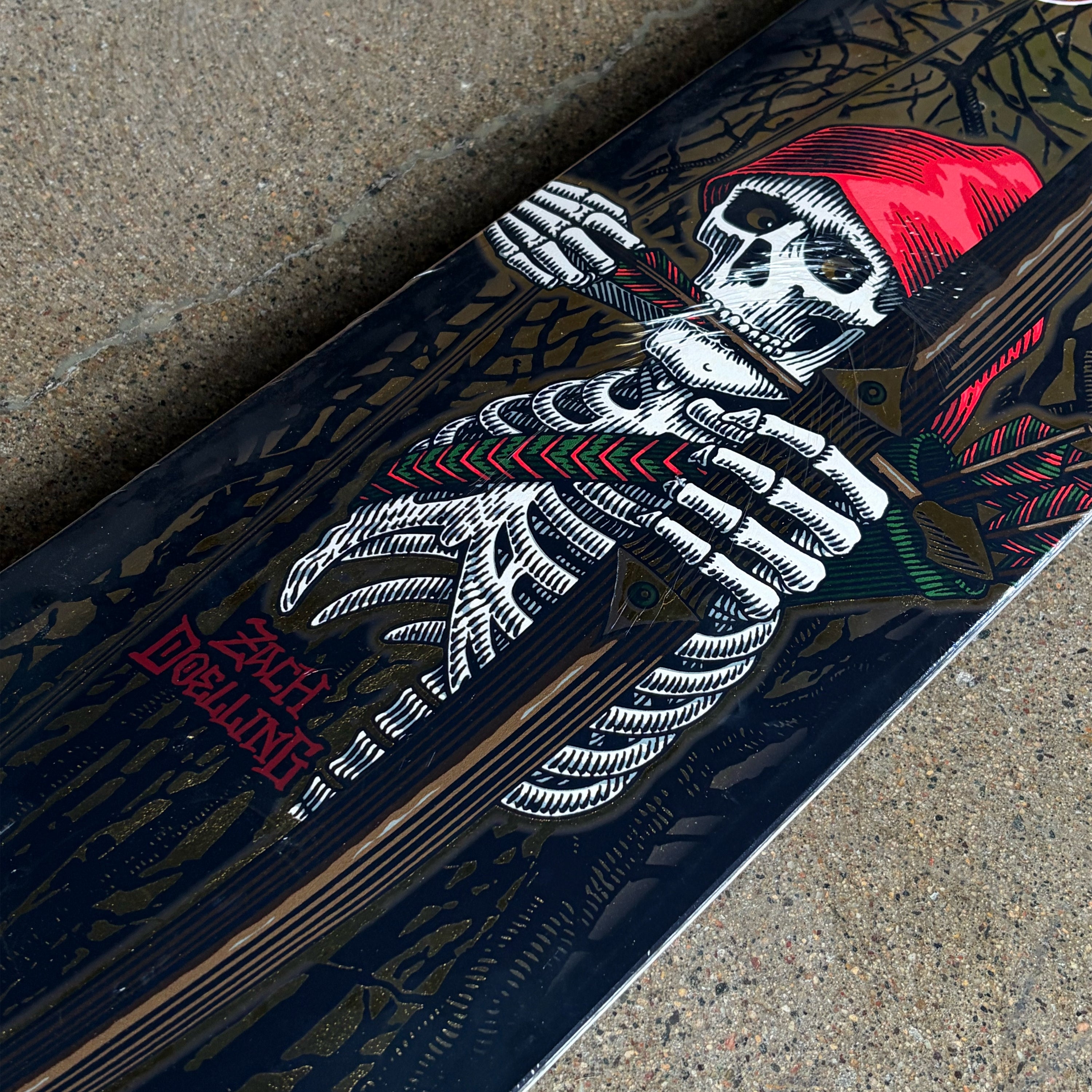 A close-up of a skateboard deck featuring a detailed graphic of a skeleton wearing a red hat, visible on a textured concrete background.