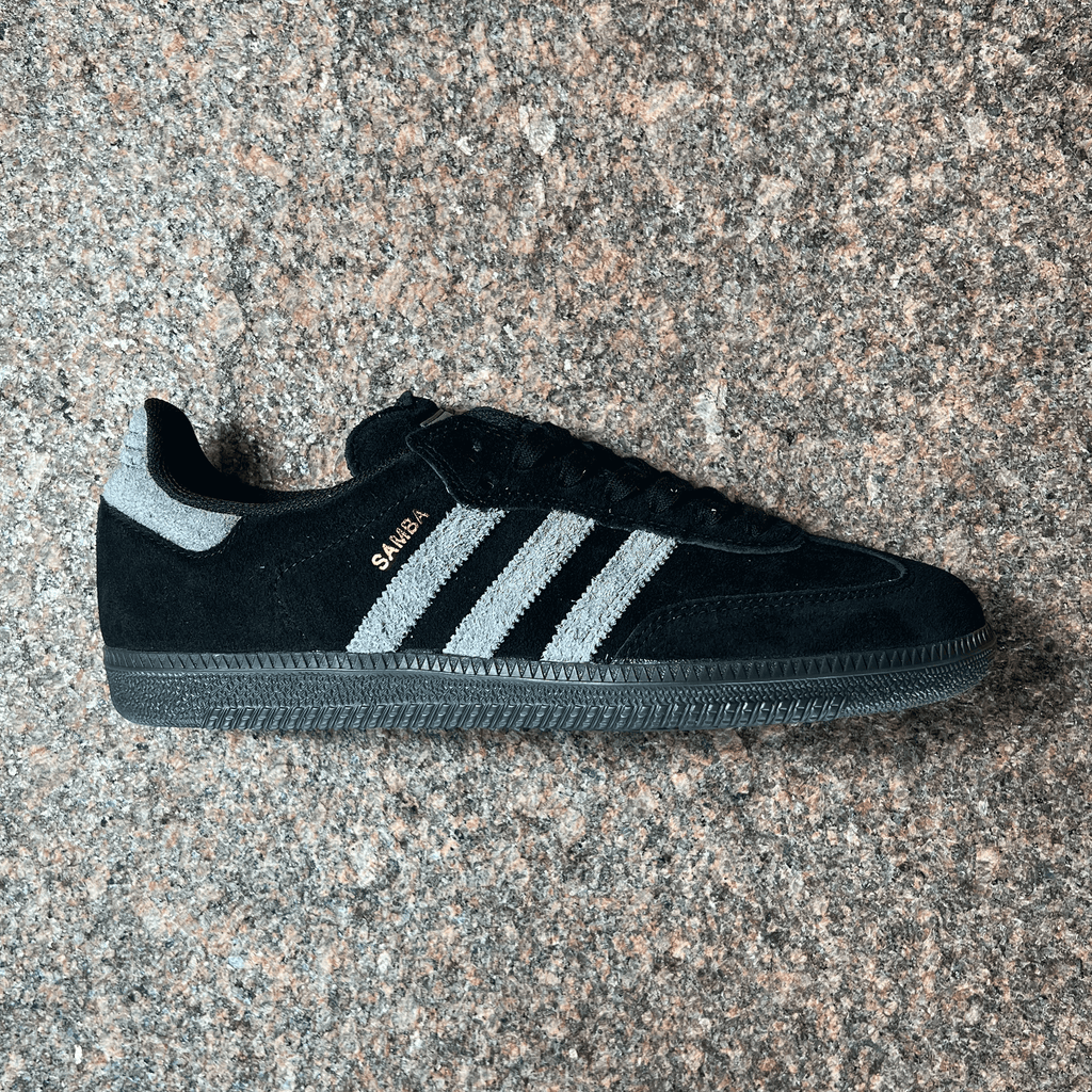 SIDE VIEW OF THE ADIDAS SAMBA ADV IN BLACK SUEDE AND GREY STRIPES, HEEL TAB. BLACK CLEAR SOLE DISPLAYED ON GRANITE