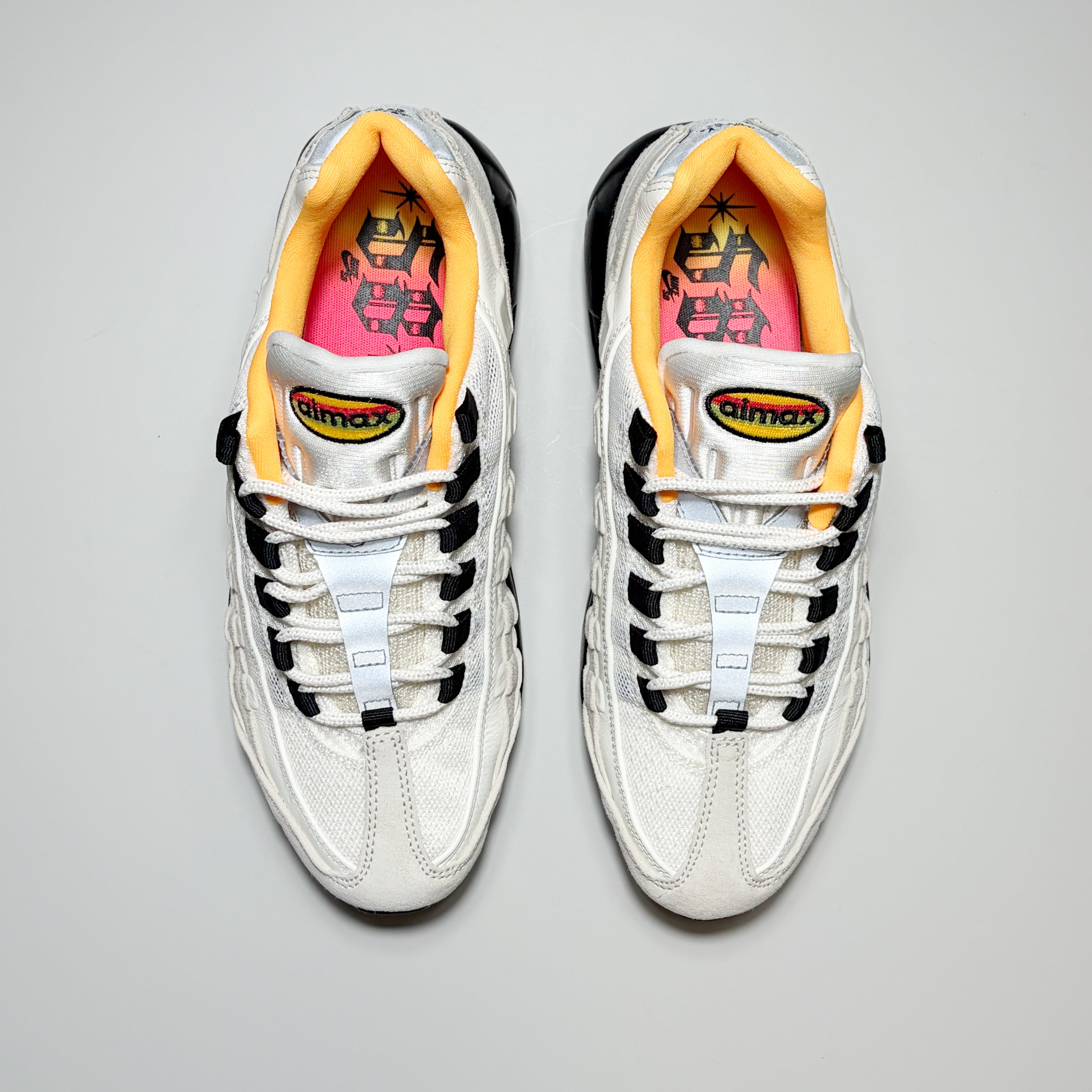 A pair of white Nike SB Air Max sneakers with black and white laces, a bright yellow collar lining, and a pink inner sole, shown from a top-down view on a light gray background.