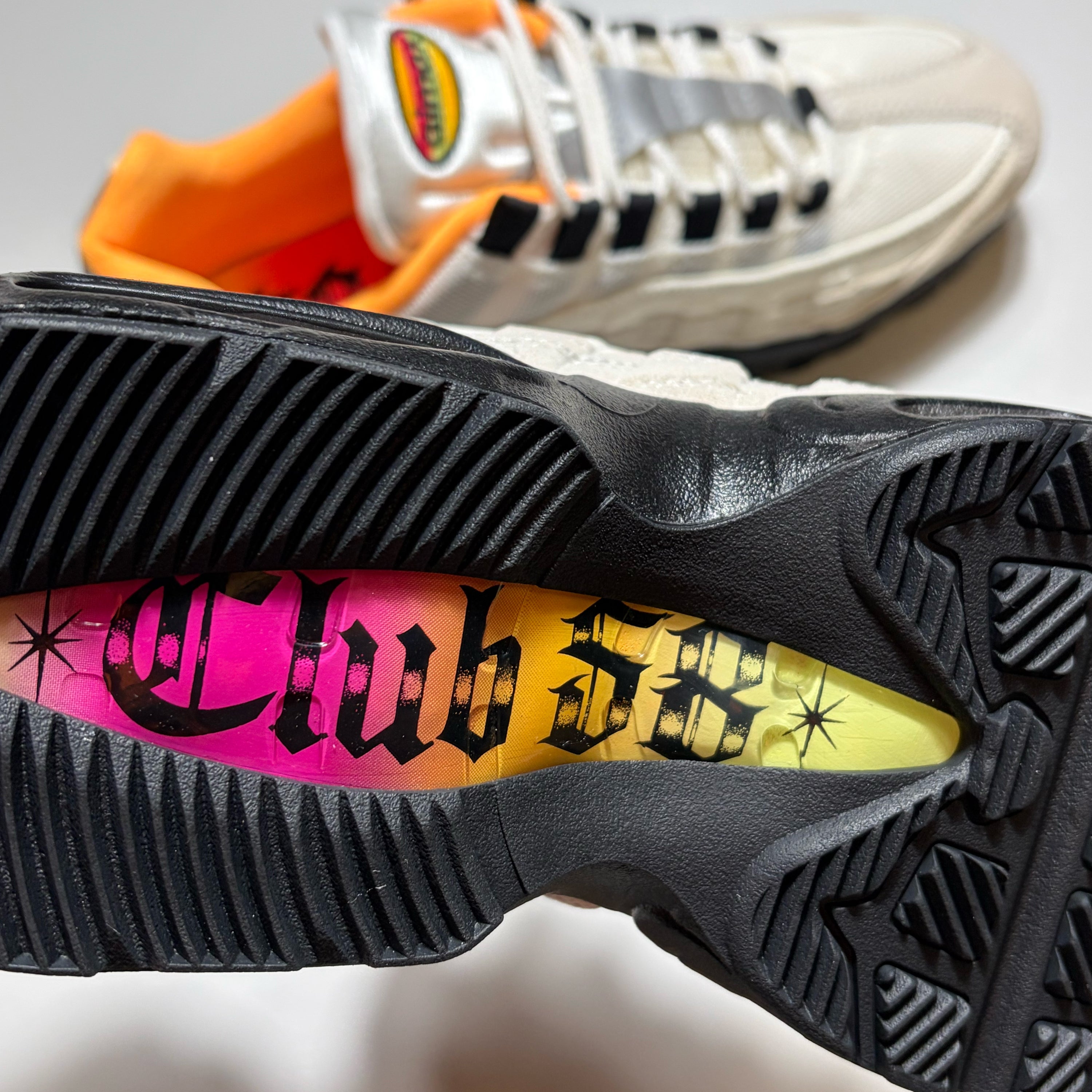 Close-up of a black athletic shoe sole with a bright pink-to-yellow gradient insole featuring the text 'Club 58' in black gothic lettering, with a second shoe blurred in the background.