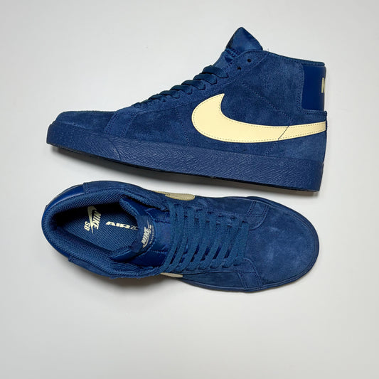 A pair of Nike SB Blazer Mid high-top sneakers in textured blue suede, featuring a light cream-colored Swoosh on the side and a matching cream heel tab, displayed on a light gray background.