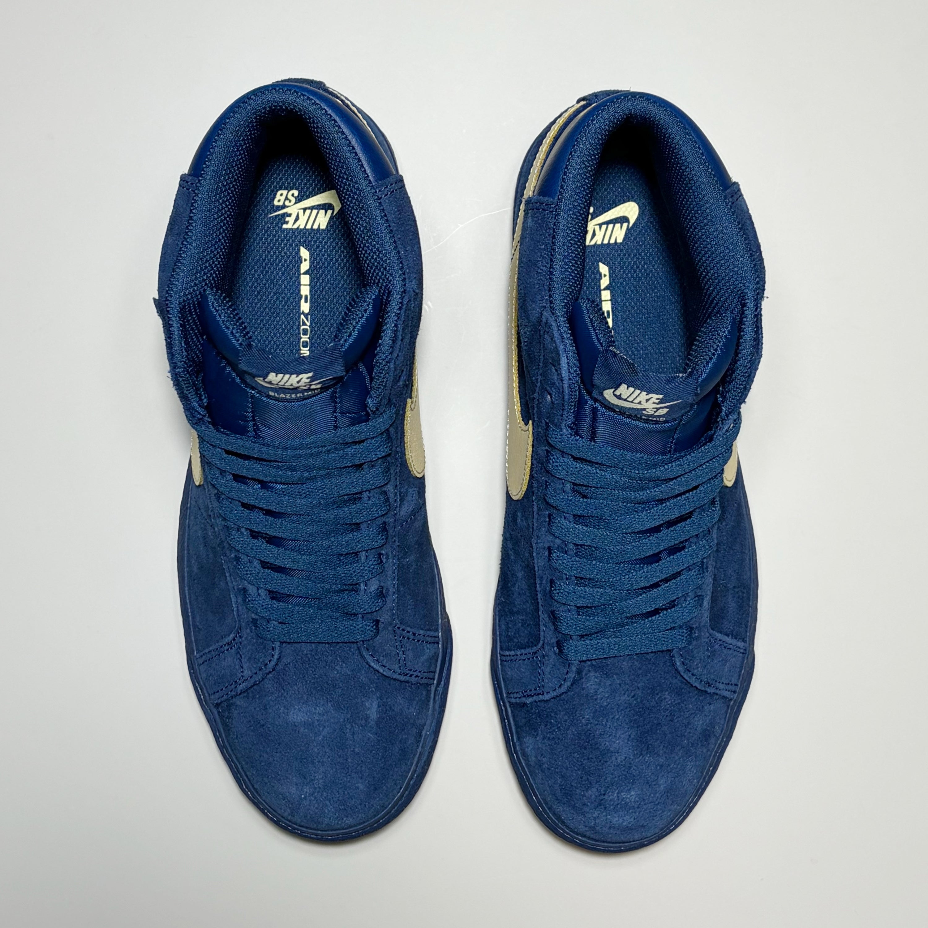 A pair of Nike SB Zoom Blazer Mid sneakers in a deep blue suede and mesh upper, featuring gold accents on the side panels and gold branding on the tongue, shown from a top-down view on a light gray background.