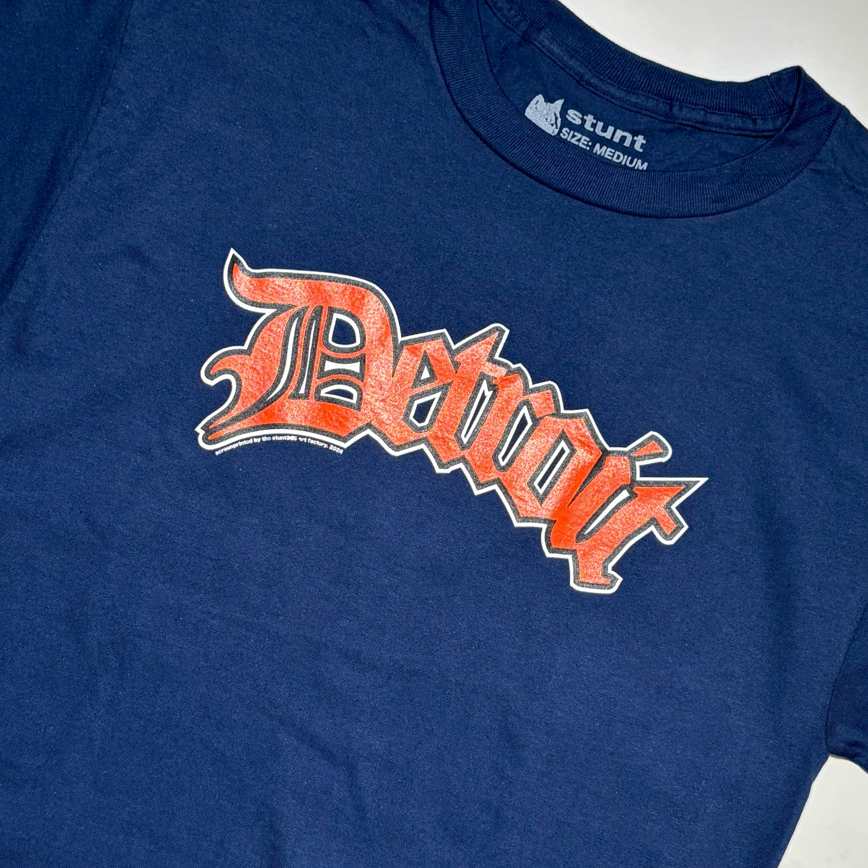 A close-up of a navy blue t-shirt featuring a distressed orange and white 'Detroit' graphic across the chest, with the 'stunt' brand logo visible inside the collar.