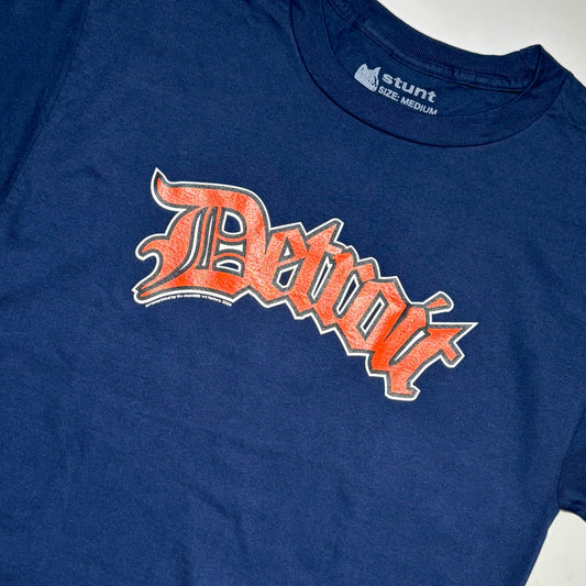 A close-up of a navy blue t-shirt featuring a distressed orange and white 'Detroit' graphic across the chest, with the 'stunt' brand logo visible inside the collar.