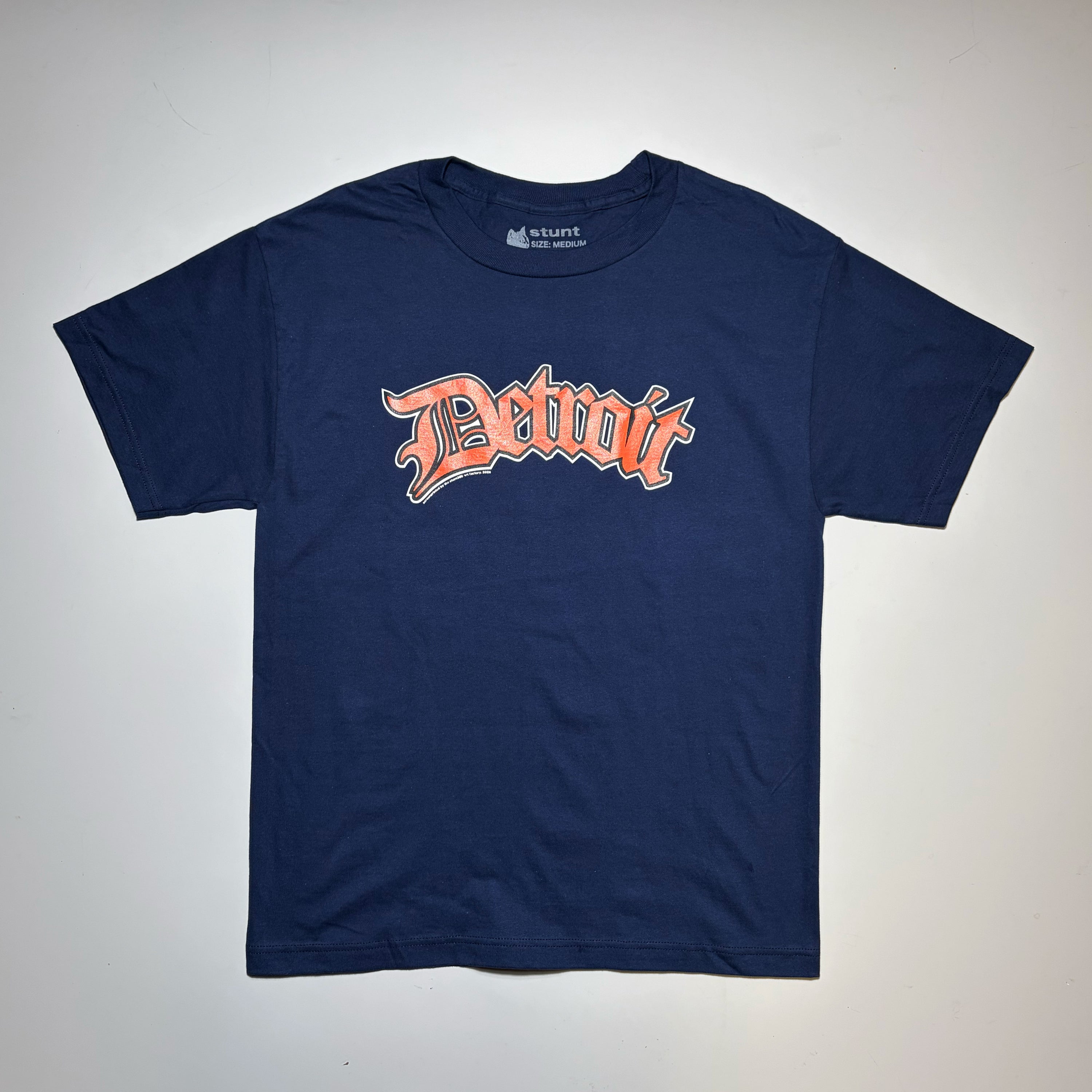 A navy blue short-sleeve t-shirt with a stylized orange and white 'Detroit' graphic across the chest, laid flat against a light gray background.