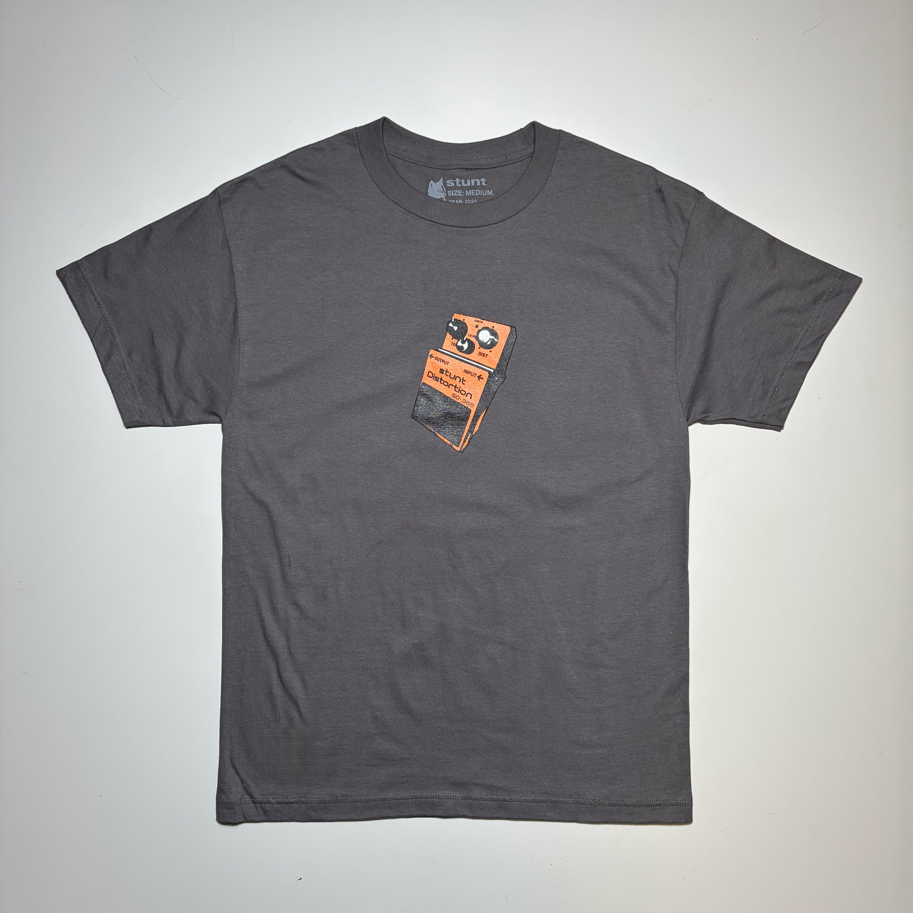 A dark gray crewneck t-shirt featuring a graphic print of an orange and black guitar effects pedal on the center chest, laid flat against a light gray background.