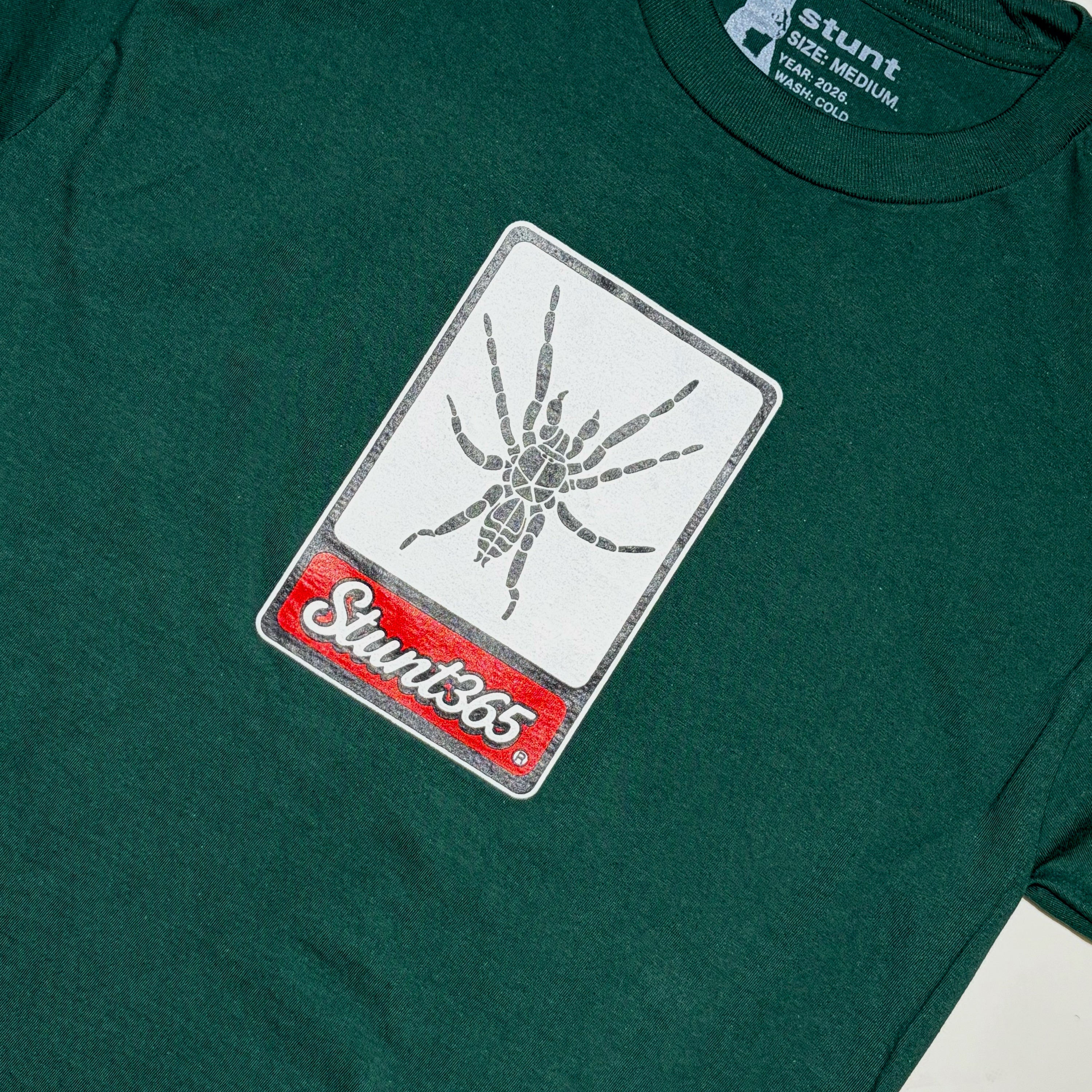 A close-up of a dark green t-shirt featuring a rectangular graphic print on the chest. The print has a white background with a gray spider illustration and a red banner at the bottom with the text 'Stunt365'.