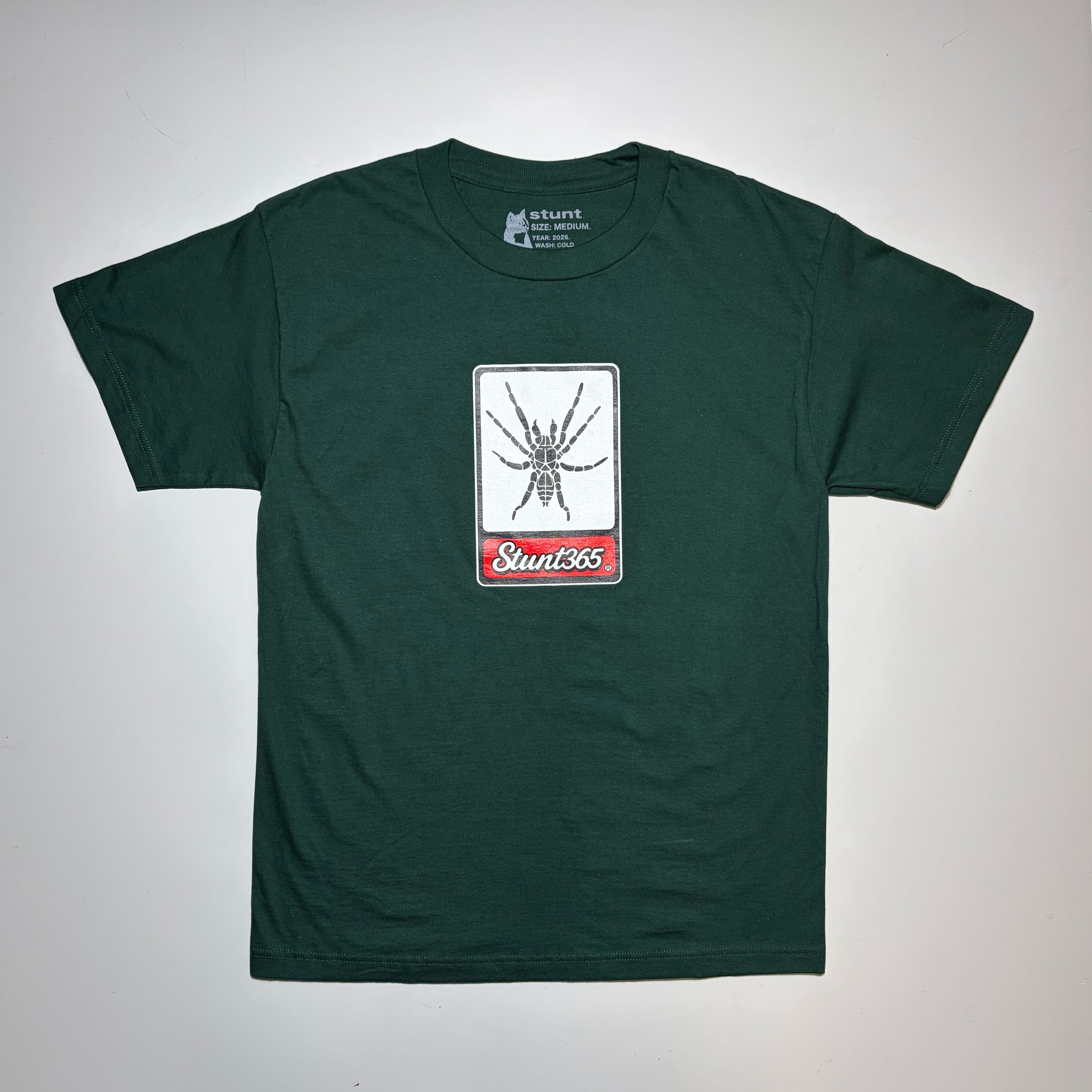 A dark green short-sleeve t-shirt laid flat on a light gray background. The center of the chest features a rectangular graphic with a black spider on a white background and a red banner below with the text 'Sunt365'.