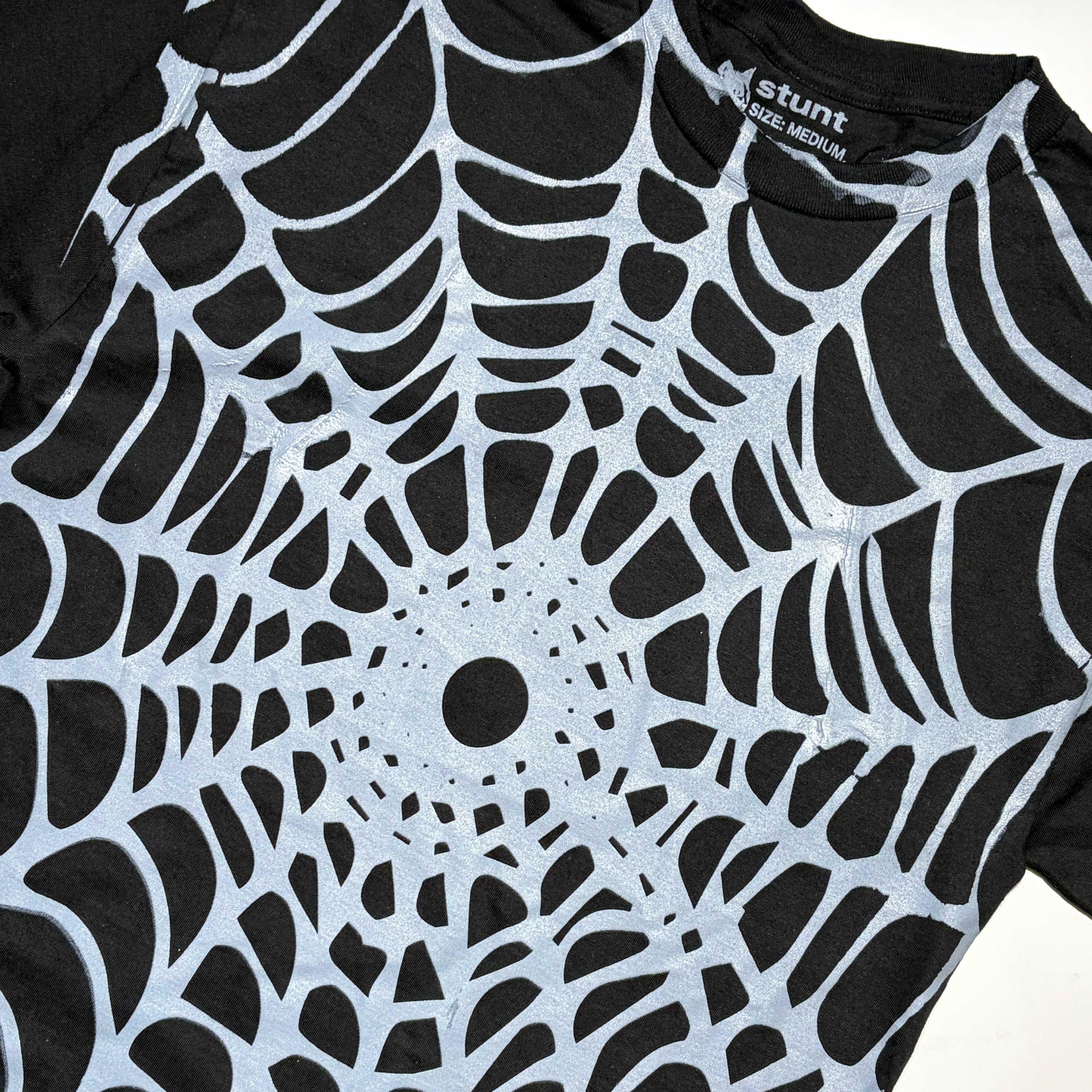 A close-up of a black short-sleeve T-shirt with a large white spiderweb pattern printed across the front. The inside collar shows the brand name 'stunt' and 'RAGE MEDIUM'.