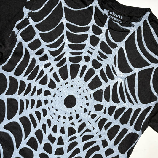 A close-up of a black short-sleeve T-shirt with a large white spiderweb pattern printed across the front. The inside collar shows the brand name 'stunt' and 'RAGE MEDIUM'.