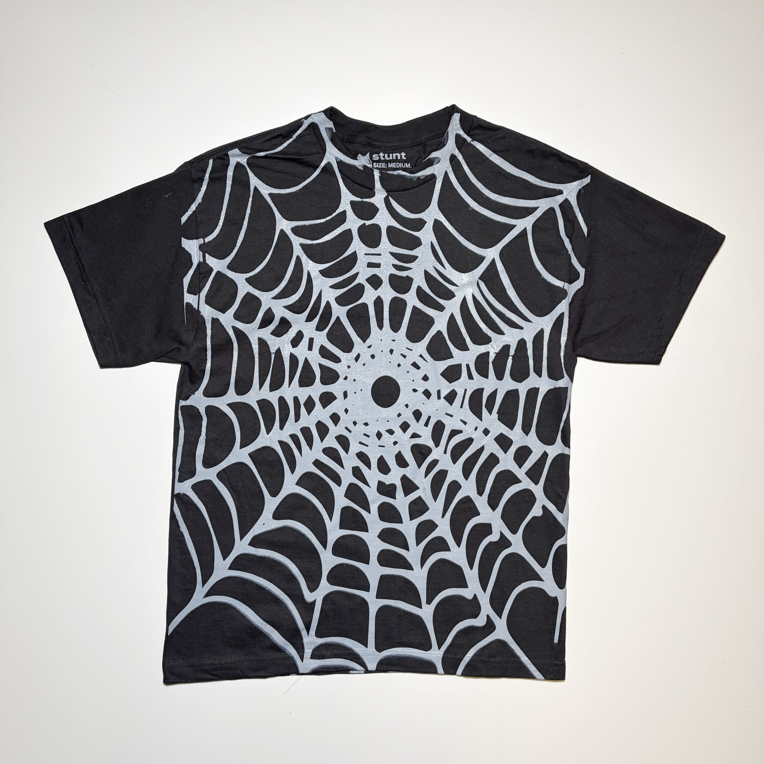 A black short-sleeve t-shirt with a large white spiderweb pattern printed across the front, laid flat against a light off-white background.