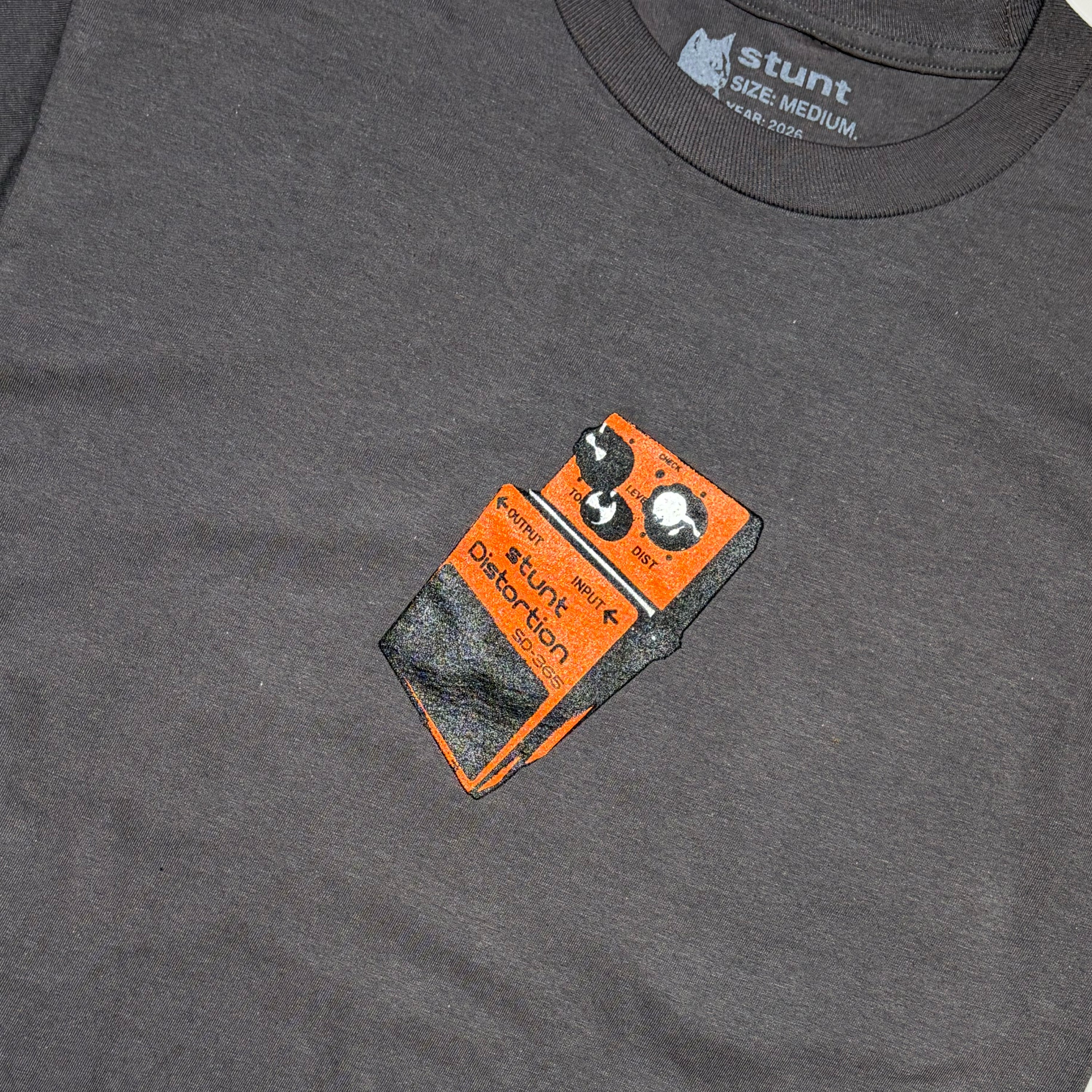 A close-up of a dark heather gray t-shirt featuring a graphic print of an orange and black Stunt Distortion guitar pedal on the chest. 