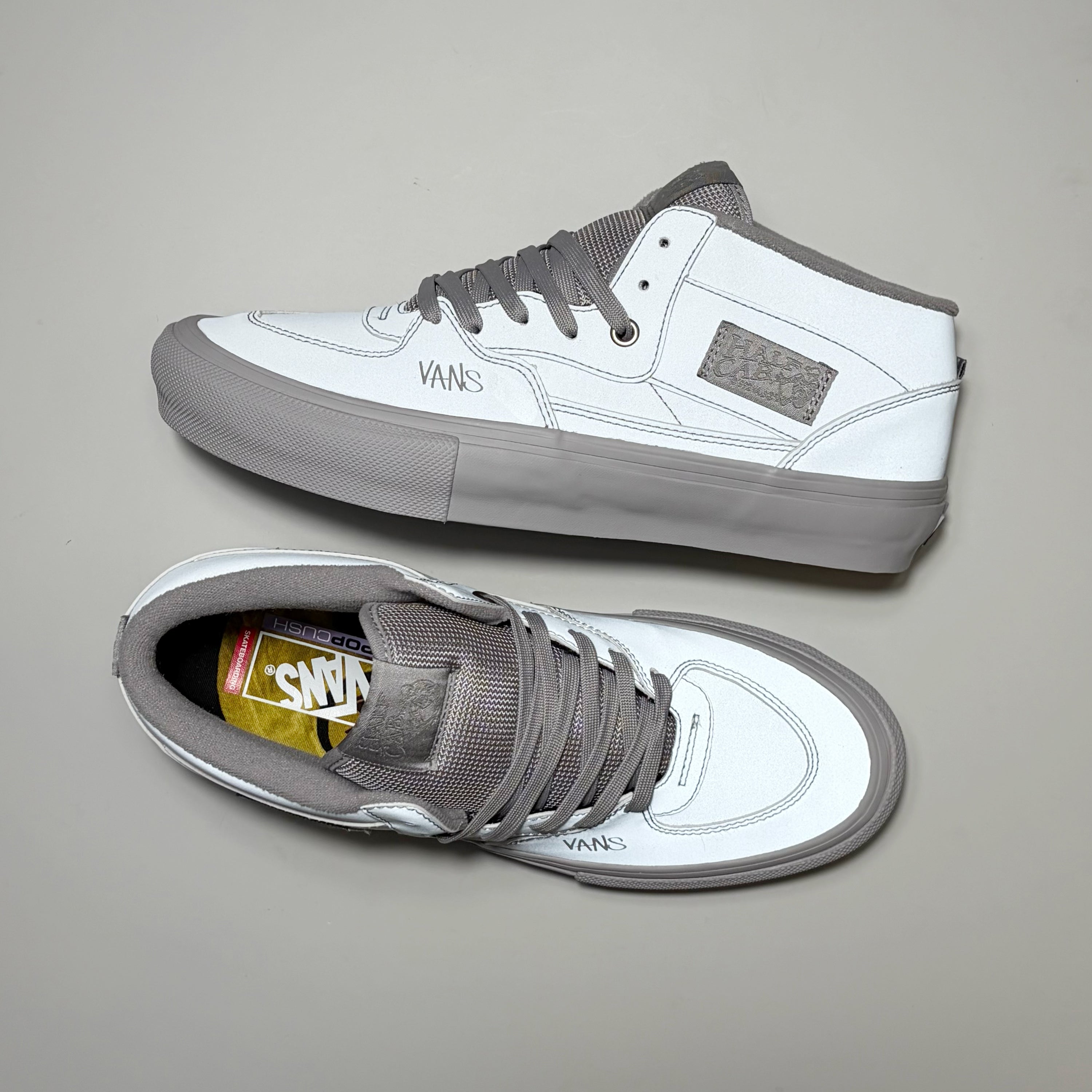A pair of Vans Skate Half Cab mid-top sneakers in white and gray, shown from both the side and top-down views on a neutral gray background. 3M reflective material by Atiba Jefferson
