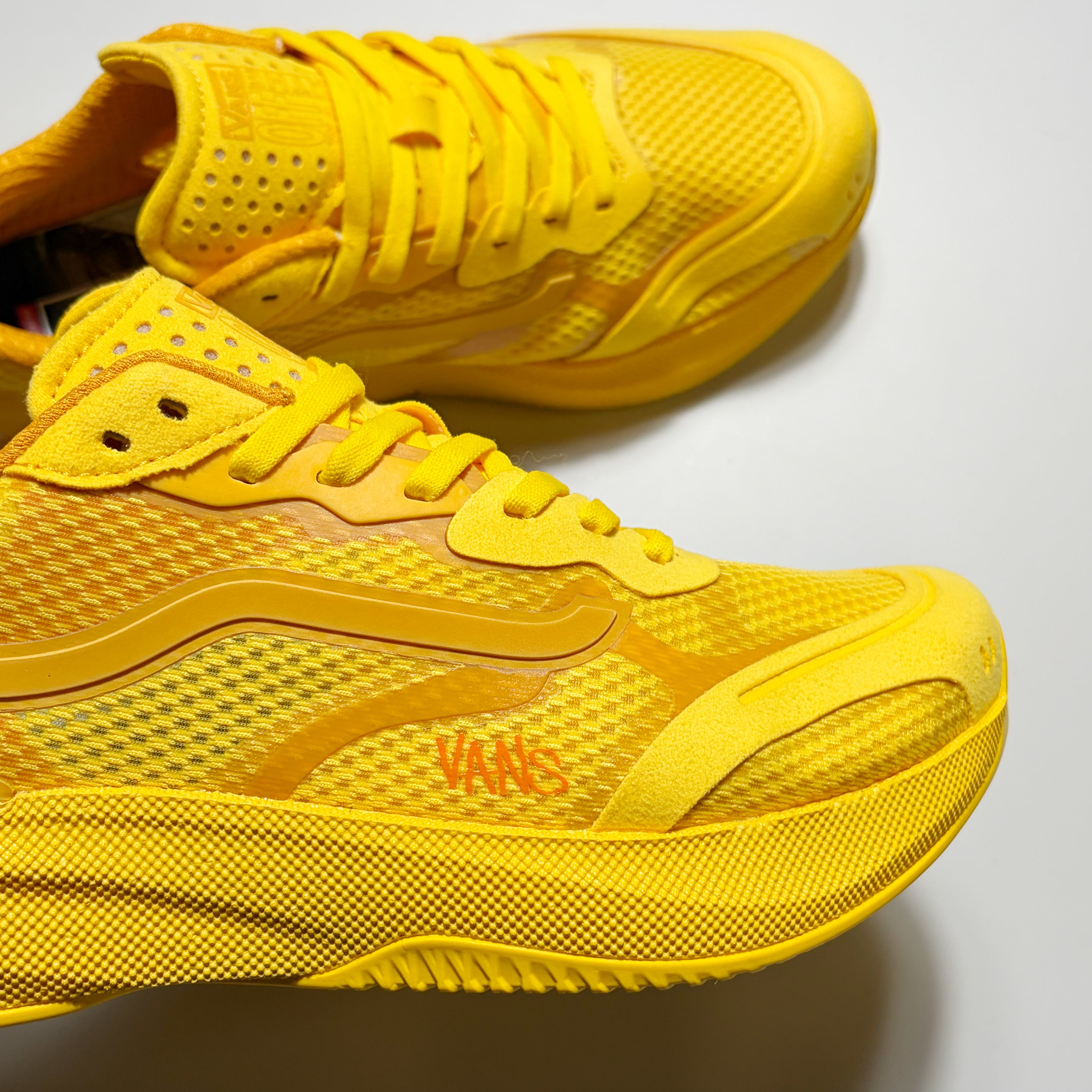 A pair of yellow Vans sneakers with a textured mesh upper and solid synthetic overlays, featuring the orange Vans logo on the side, set against a plain light gray background.