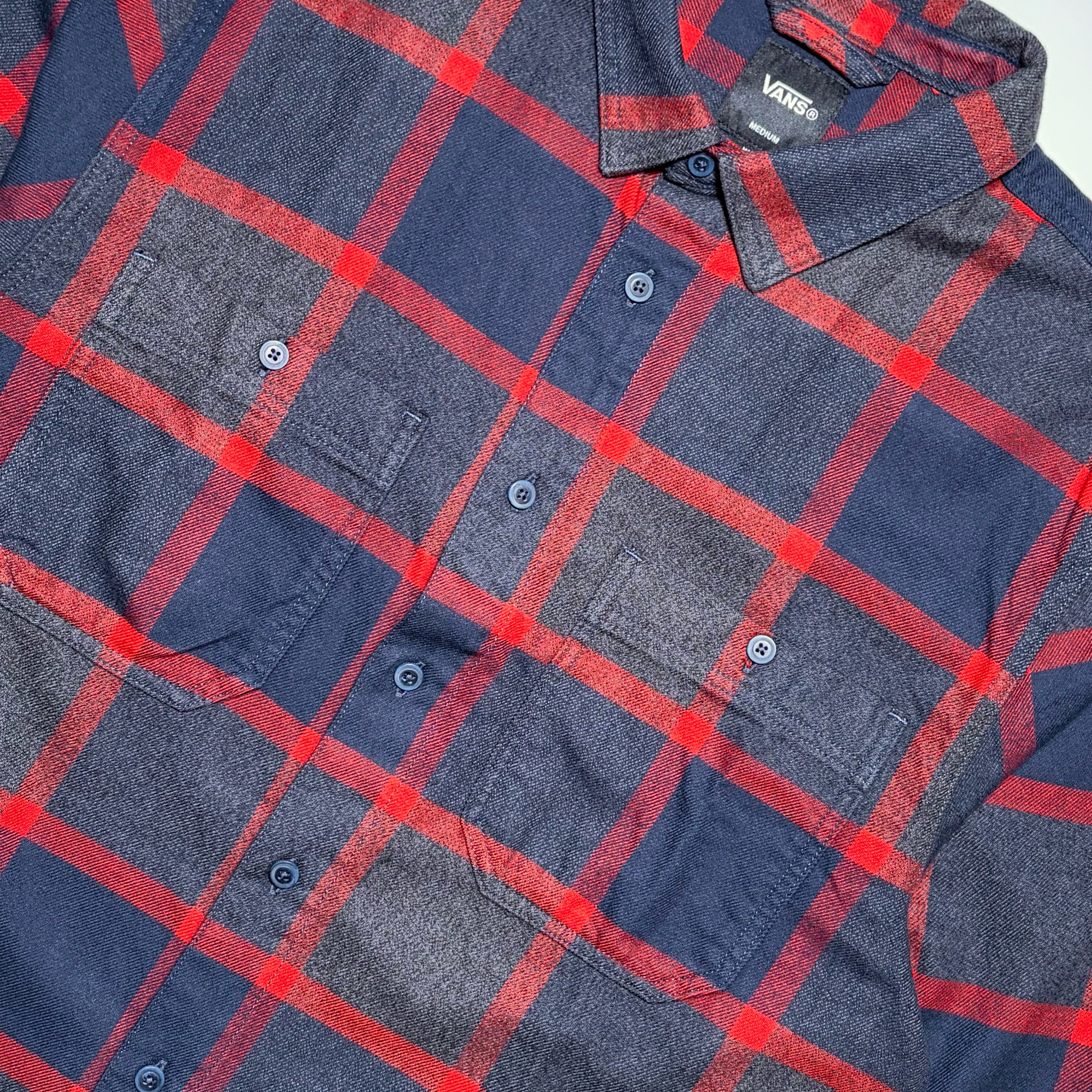 A close-up of a Vans plaid button-up shirt in navy blue and red, featuring two chest pockets and a visible Vans neck label.