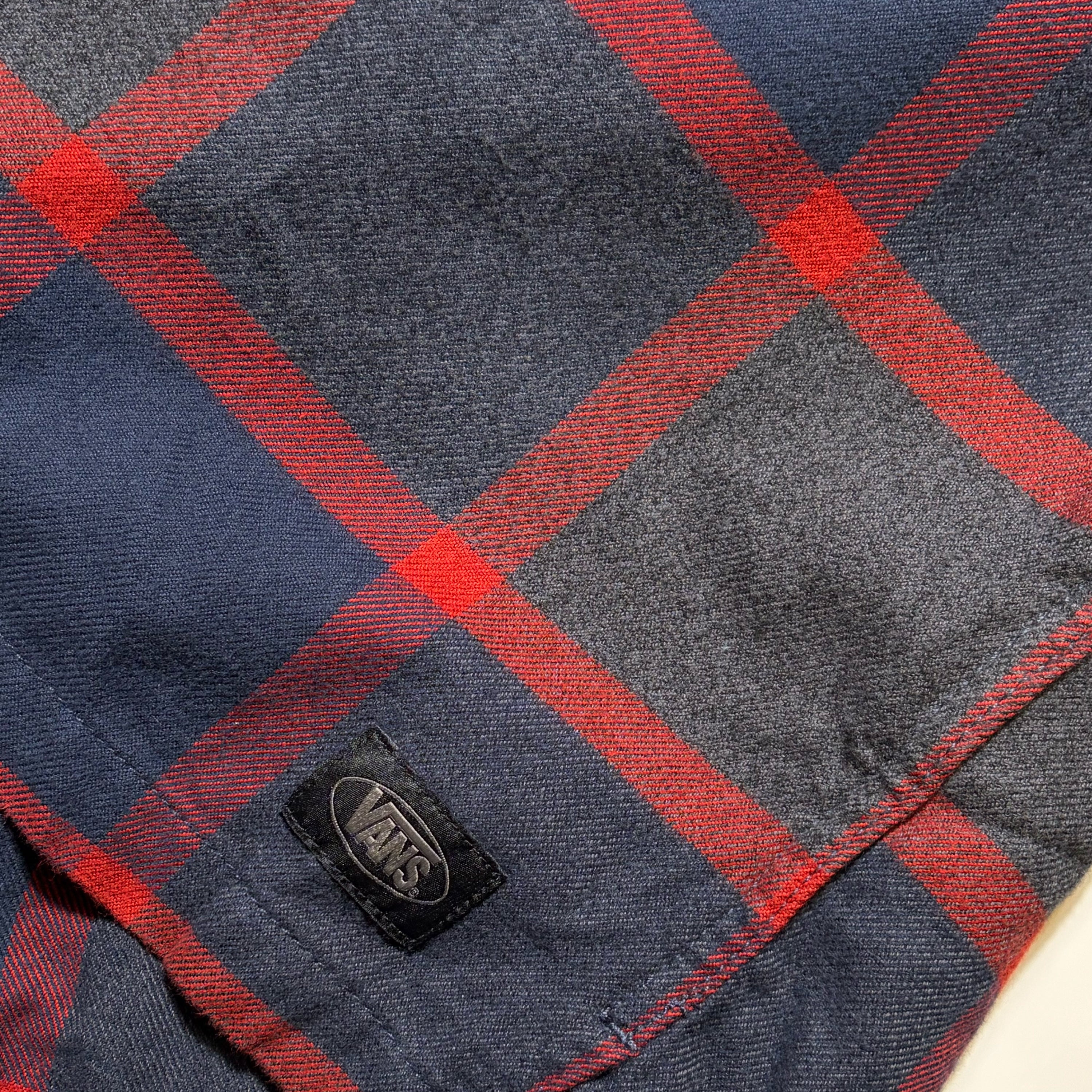 Close-up of a Vans plaid fabric in navy blue and red, showing a black Vans logo patch sewn onto the side.