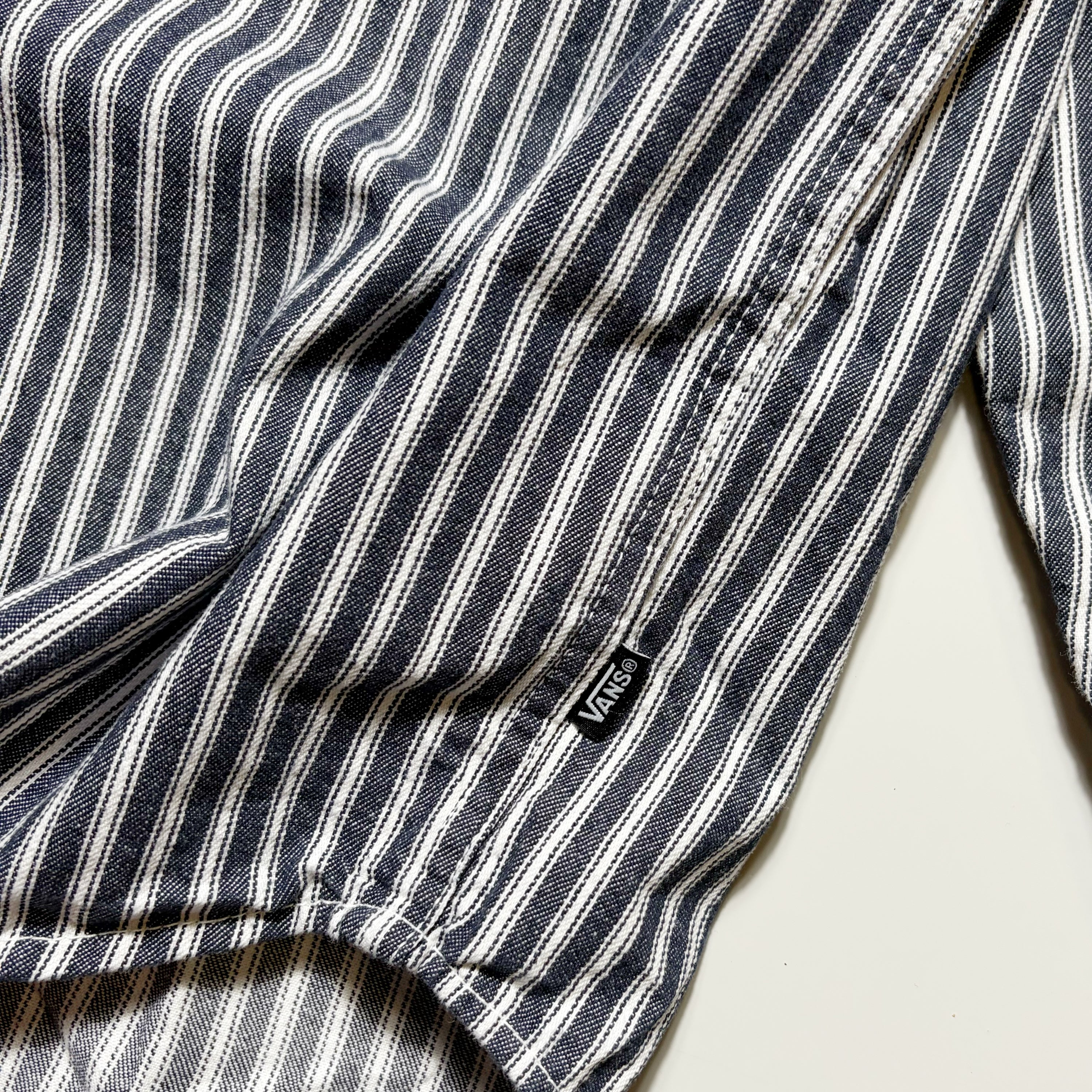 Close-up of a striped garment, likely a shirt or jacket sleeve, featuring vertical navy, white, and gray stripes with a small black Vans logo tag.