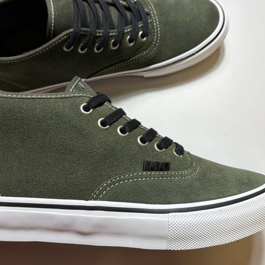 An olive green suede low-top sneaker with black laces, black trim, and a white rubber sole, shown alongside a second shoe in the background.